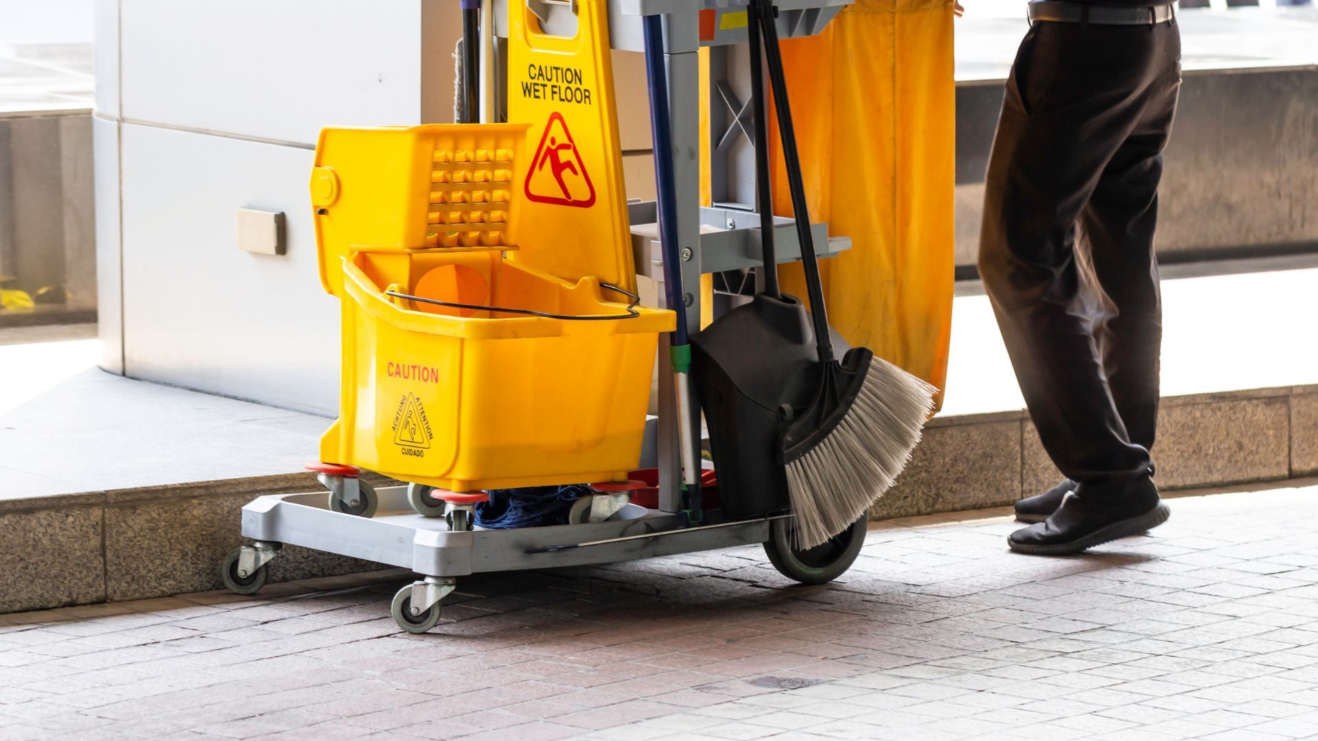 Yellow cleaning cart with mop, broom, and worker's legs.
