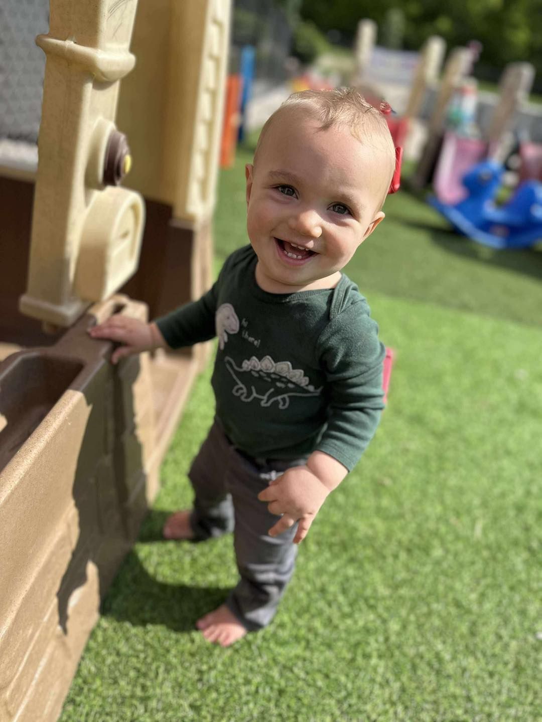 Child smiling and looking at the camera, wearing a green shirt with a dinosaur, standing on green turf near a play structure.