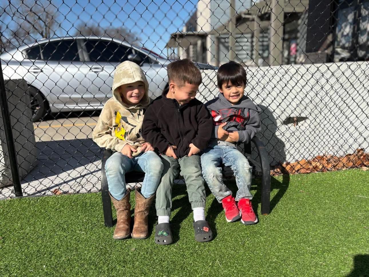 Three children seated on a bench outdoors near a fence, smiling. One in tan hoodie, jeans, boots;