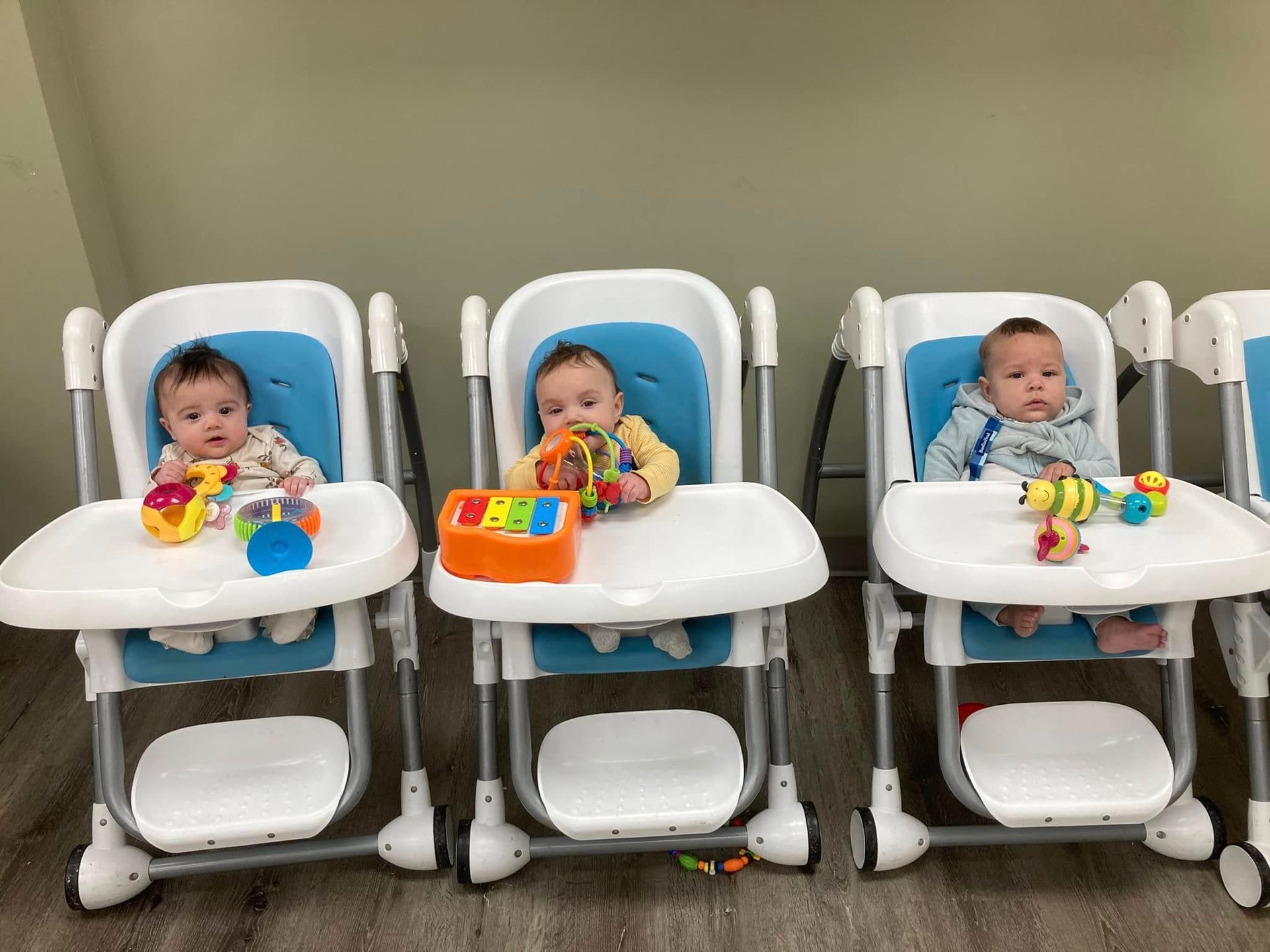 Three babies in high chairs with toys. They sit in a room with light blue seats and white trays.