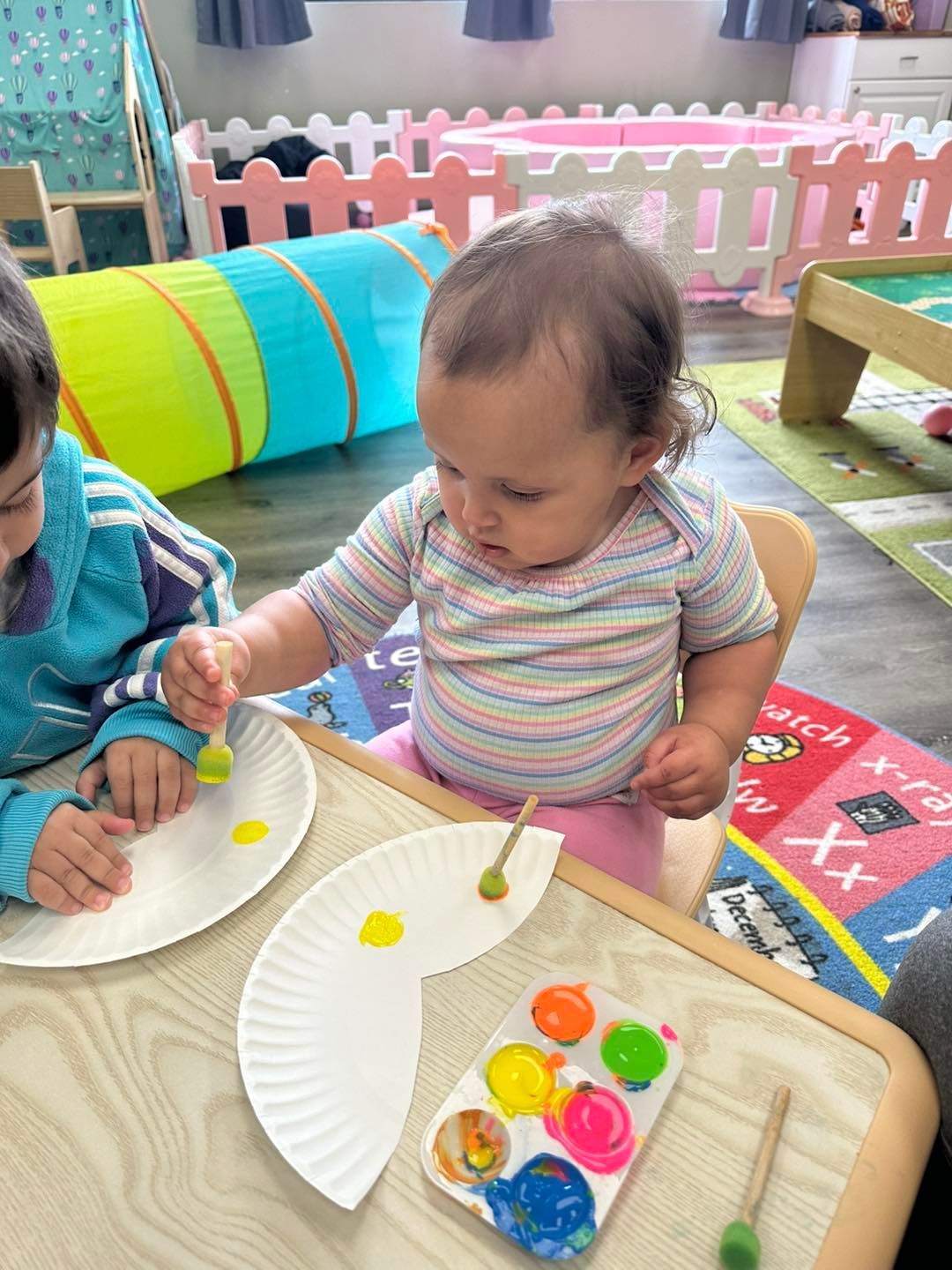 A child paints with yellow on a paper plate. Other child at the table. Paint palette on the table.