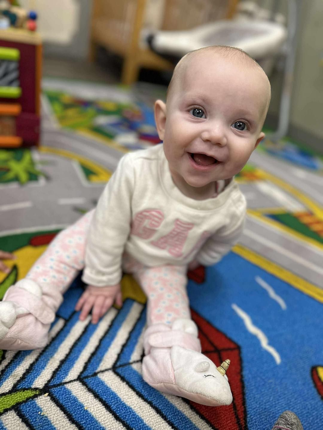 Smiling baby wearing pink and white clothes sits on a colorful rug.