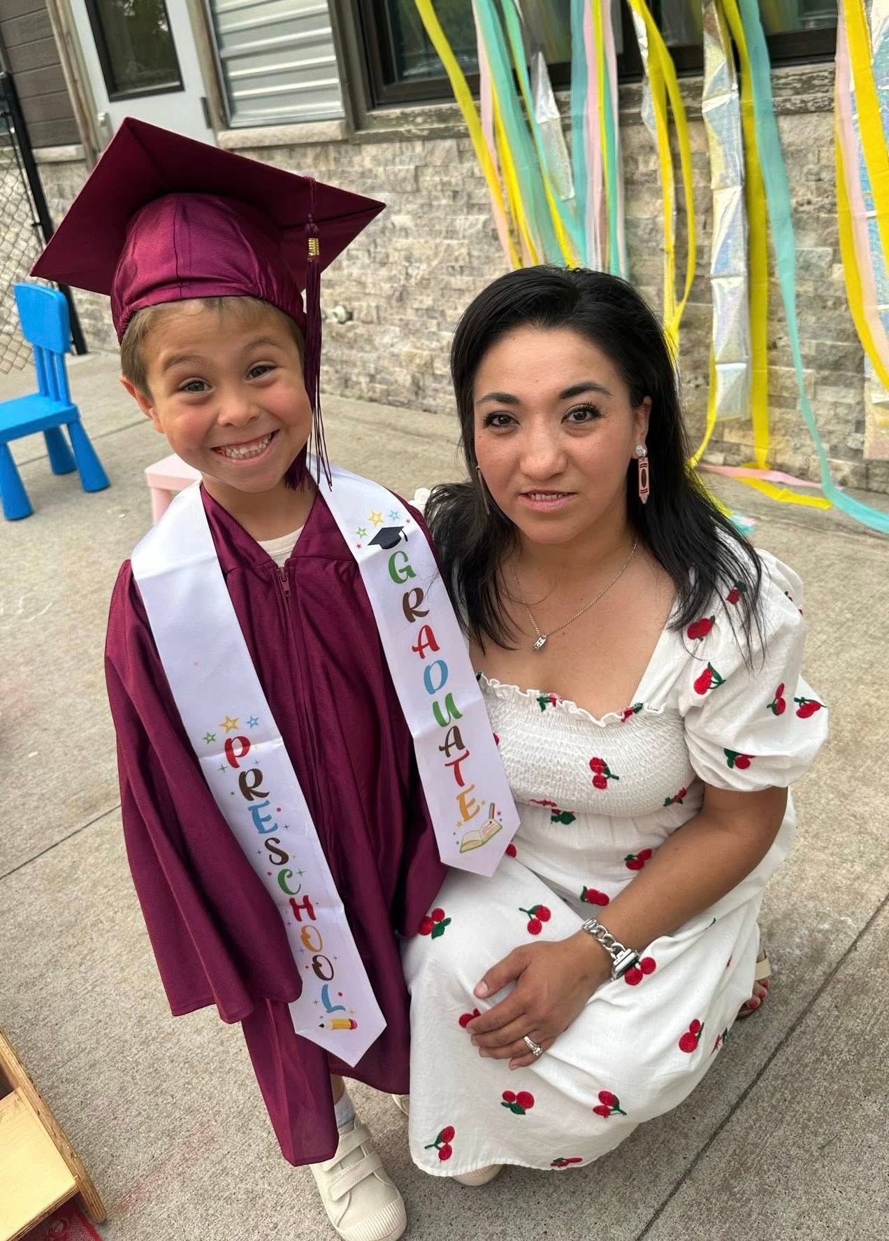 A child in a graduation gown and sash poses with a woman, outdoors.