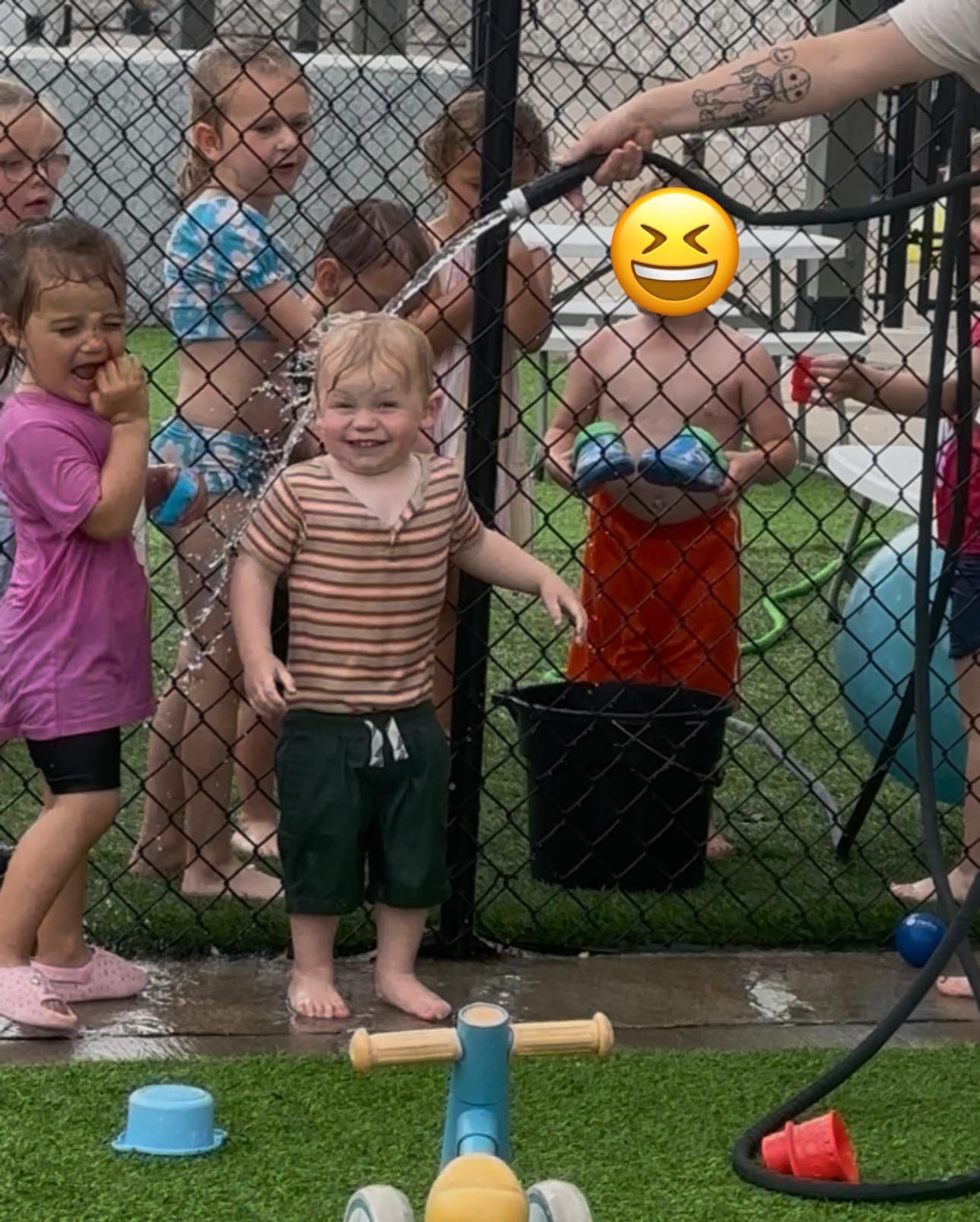 Children playing with water from a hose outside. Some are smiling, and one has an emoji over their face.