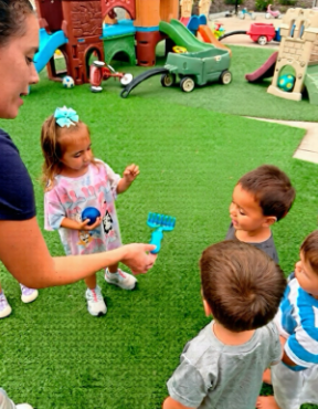Children and an adult playing outdoors with toys on green turf.