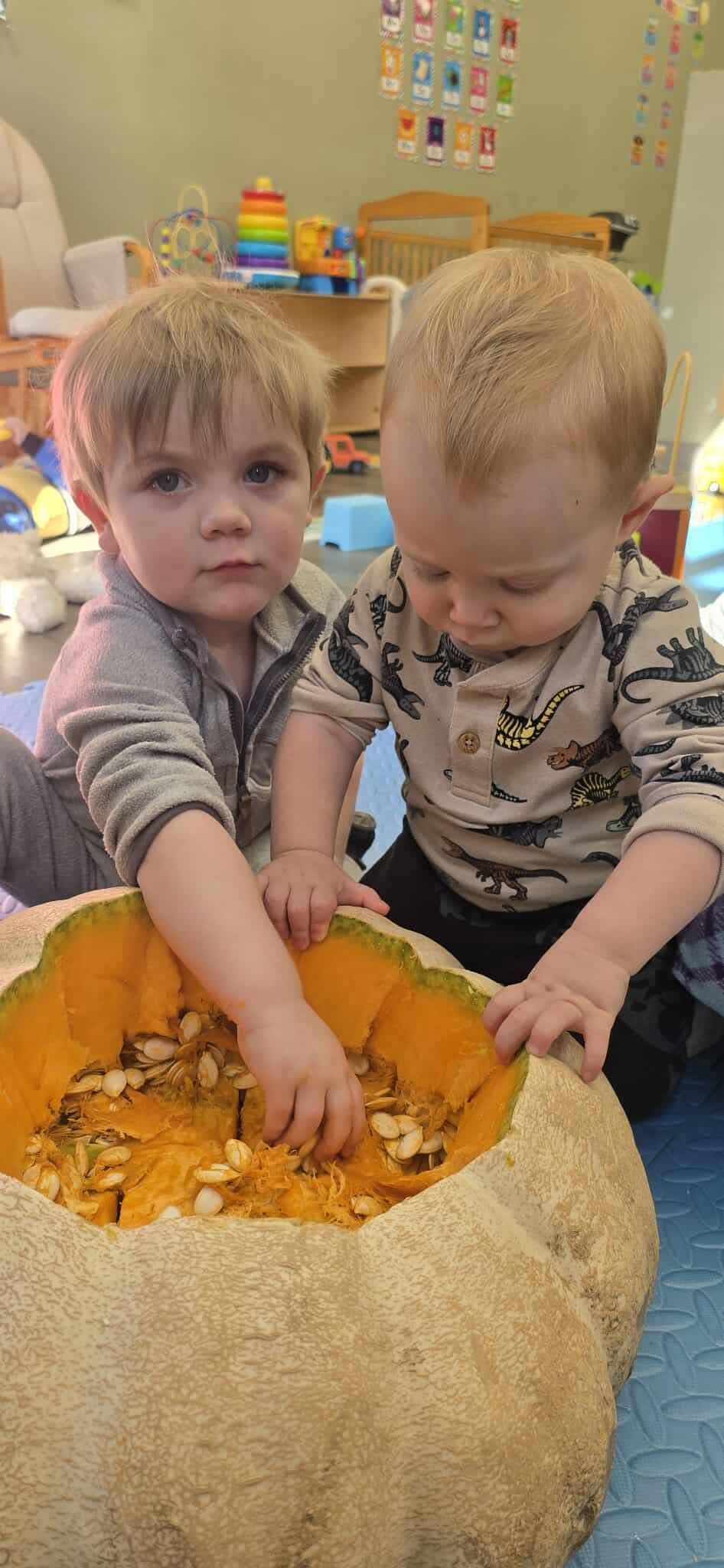 Two toddlers in a classroom paint with smiles. One has a blue handprint.