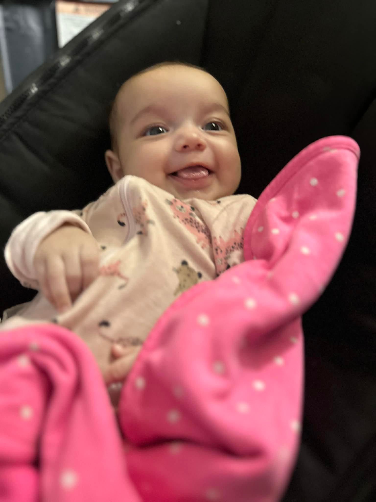 Smiling baby wearing pink and white clothes sits on a colorful rug.