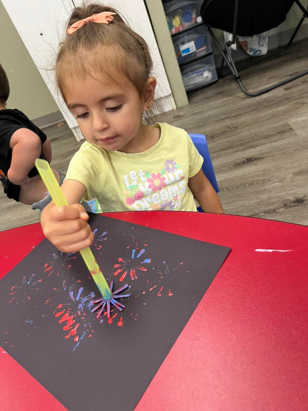 Child creating artwork using a brush, making colorful fireworks on a black paper, sitting at a red table.