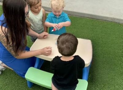An adult and three children sit around a small table on green turf, interacting with a white craft item.