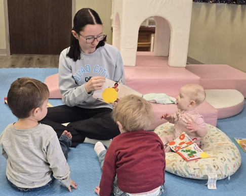 Three babies in high chairs with toys. They sit in a room with light blue seats and white trays.