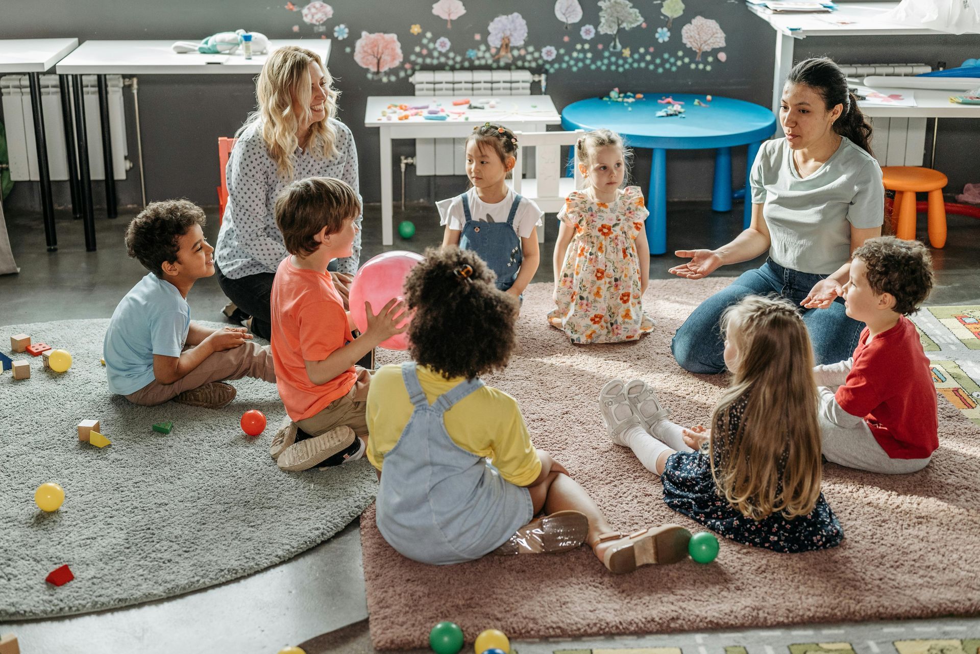 Children and two teachers sit on a rug in a classroom.