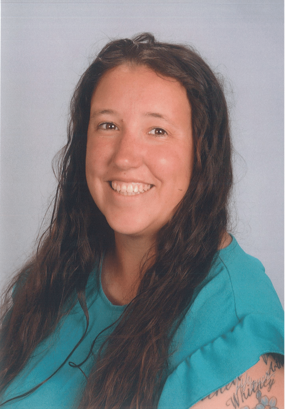 Woman with long dark hair smiles, teal shirt, arm tattoo. 