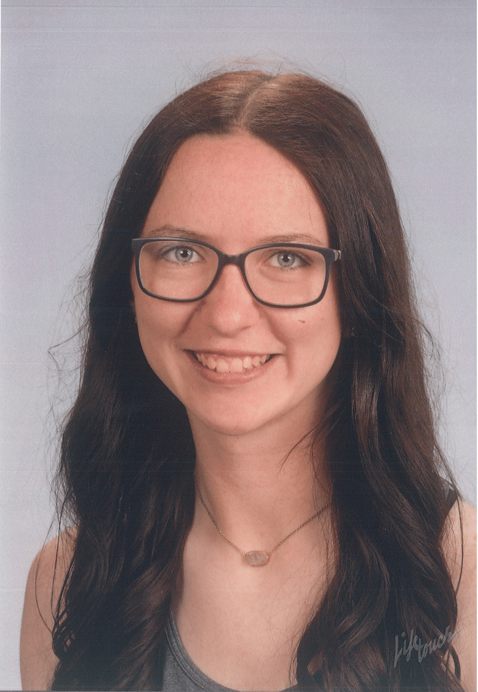 Woman with glasses smiles; dark hair, neutral background, wearing a necklace.