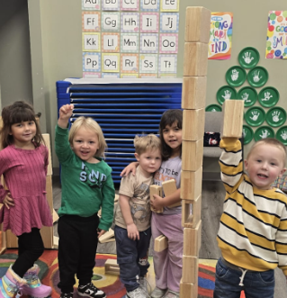 Children building a tower with blocks in a classroom. They are smiling and raising their hands.