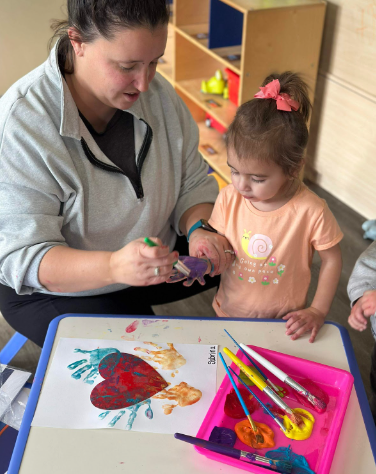 Woman assisting a child with handprint art. A red heart shape and paint supplies are on the table.