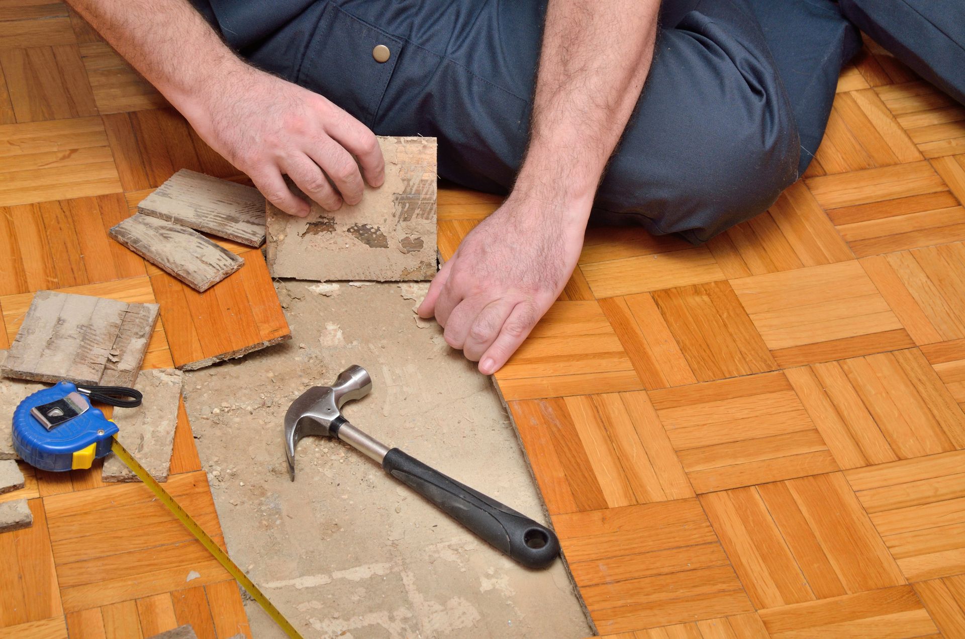 Person kneeling, repairing parquet flooring; holding a tile, hammer nearby, measuring tape.