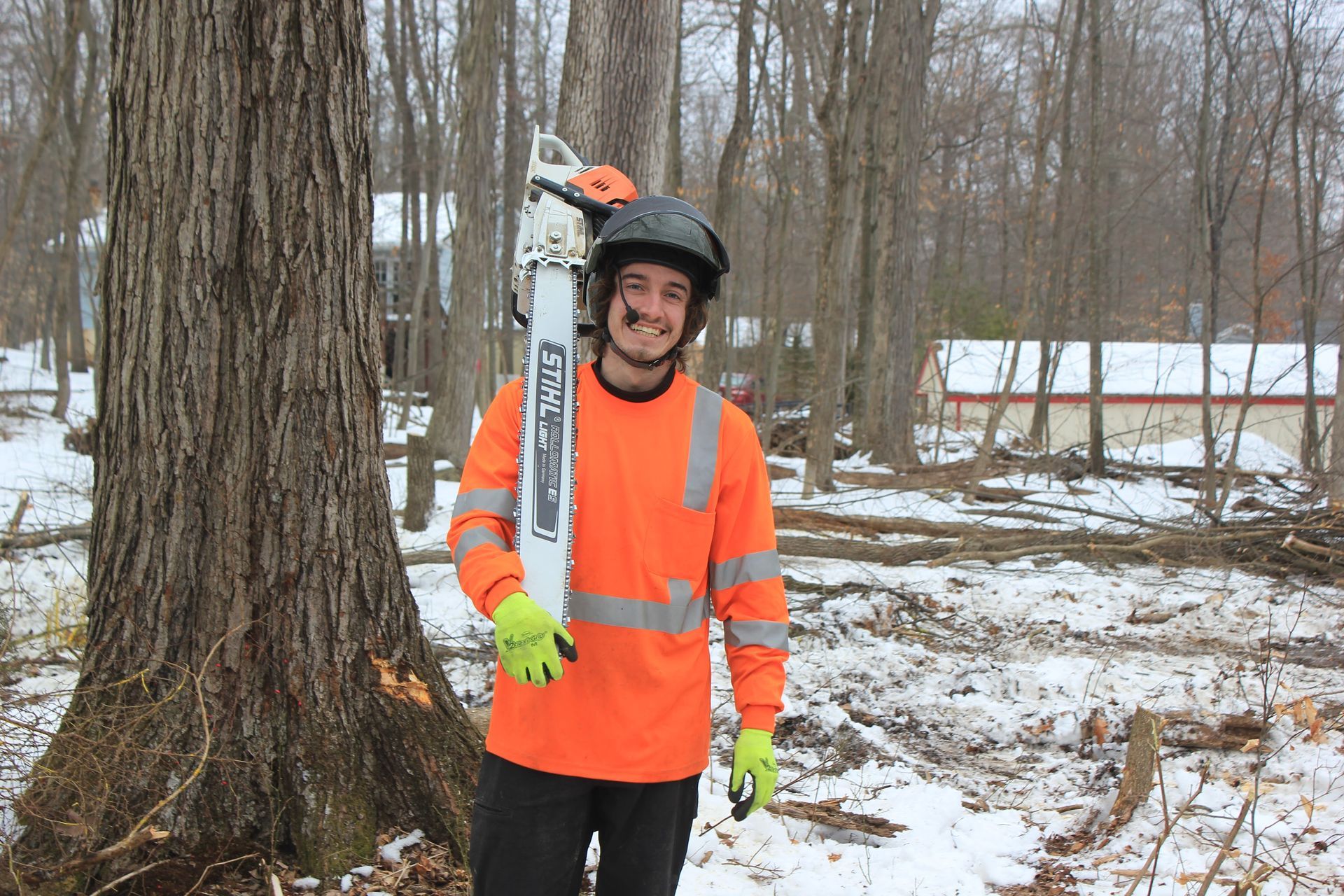 A man in an orange shirt is holding a chainsaw in a snowy forest.