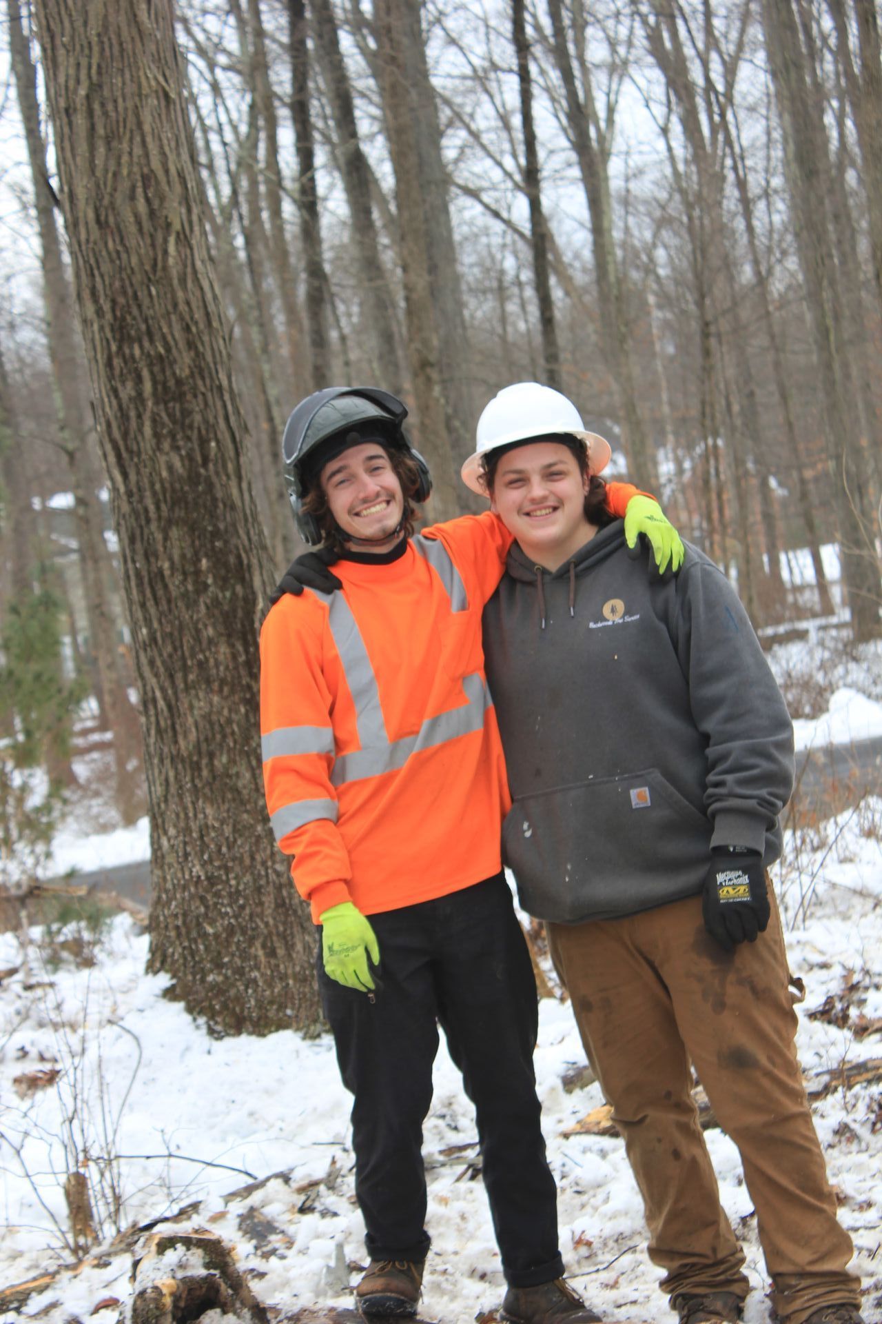 Two men are posing for a picture in the snow in front of a tree.