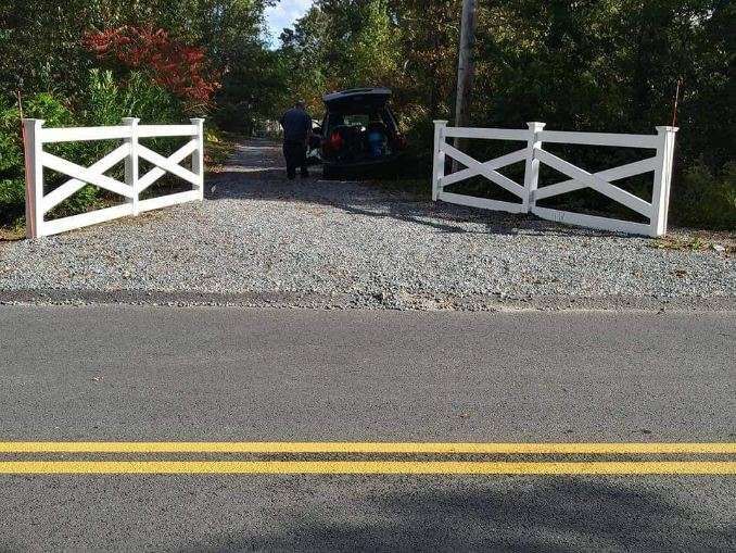 A car is parked in a gravel driveway behind a white fence.