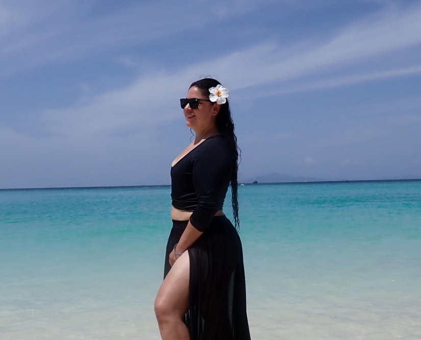A woman wearing sunglasses and a flower in her hair is standing on a beach.