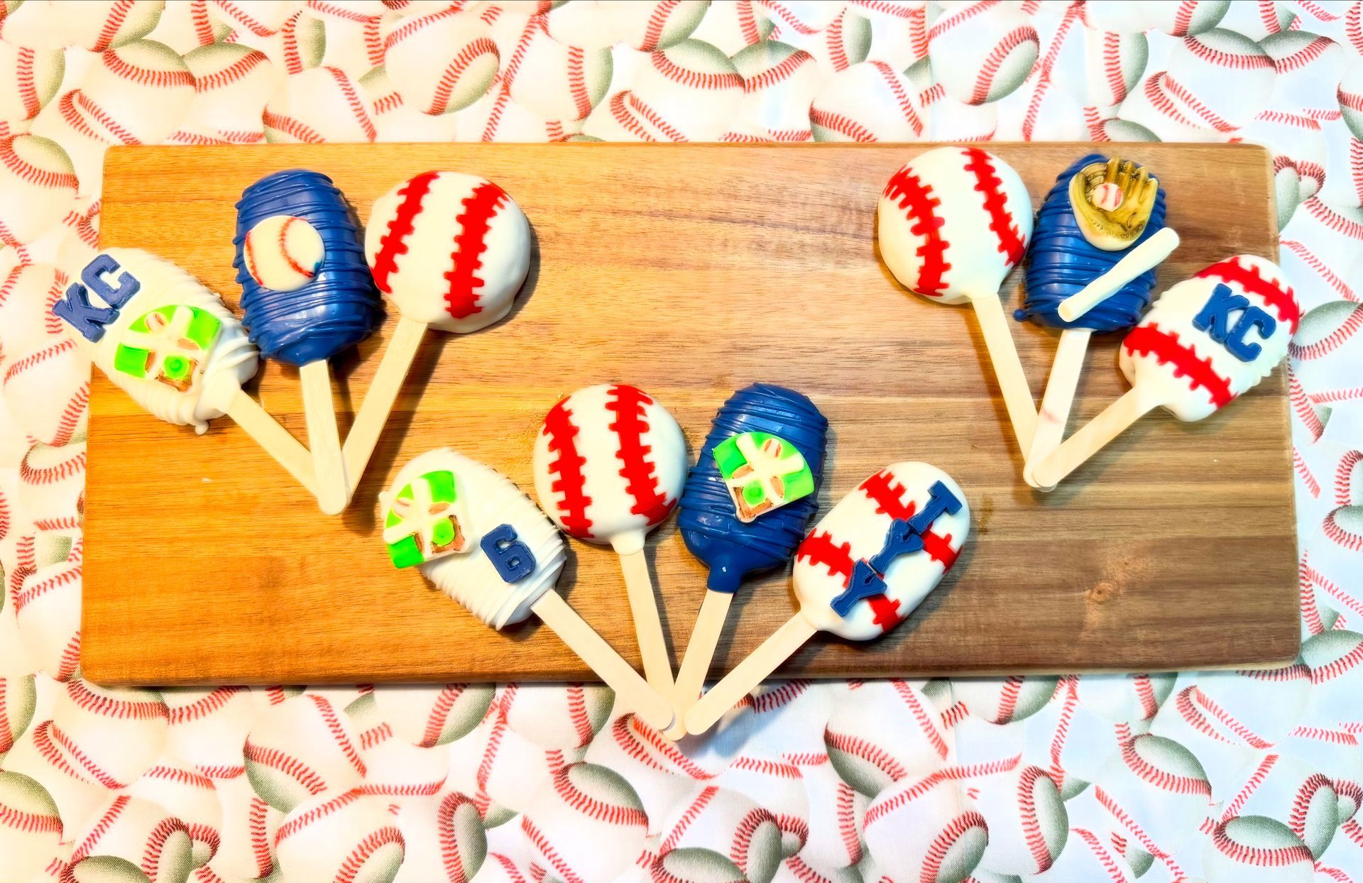 Baseball-themed cake pops on a wooden board. Features include baseball, glove, and team logo designs in red, white, blue, and green.