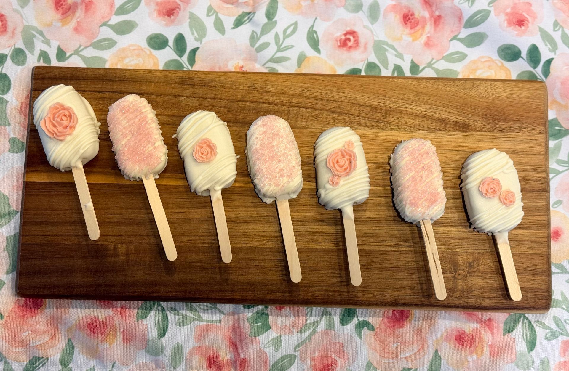 Cake pops on sticks, decorated with pink sprinkles and flowers, on a wooden board, floral background.