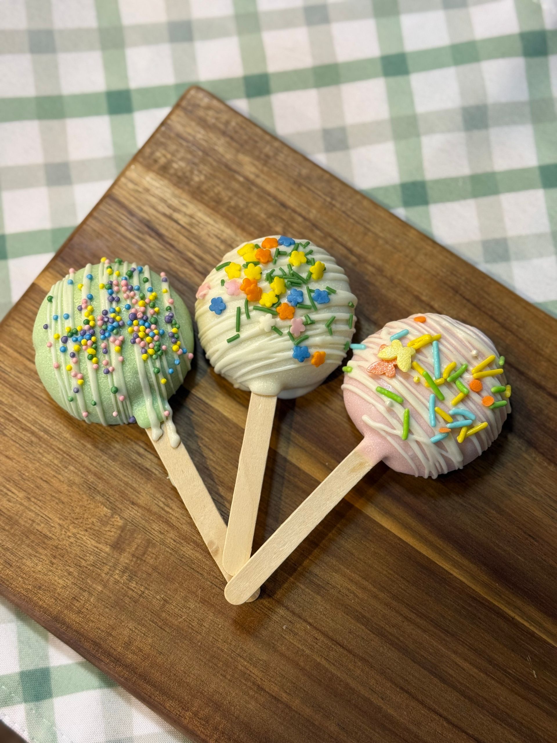 Three frosted cake pops on wooden sticks, decorated with sprinkles, on a wood board.