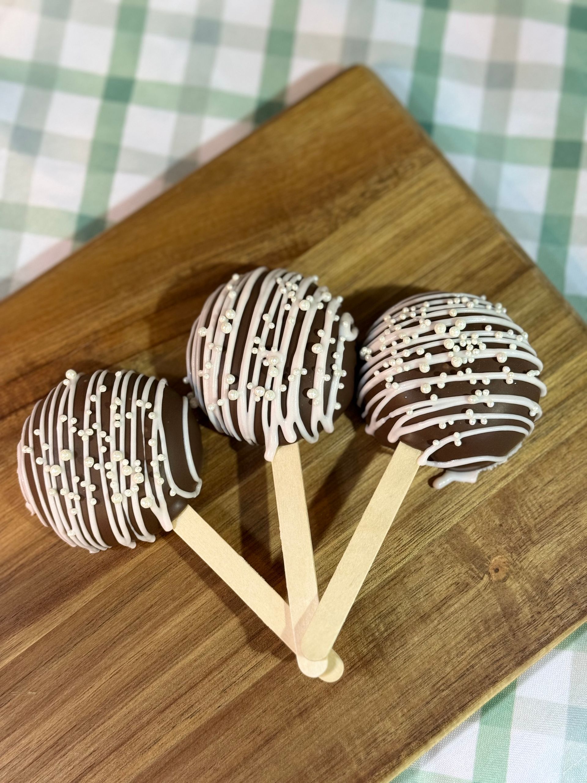 Three chocolate-covered cake pops on sticks, drizzled with white chocolate, arranged on a wooden board.