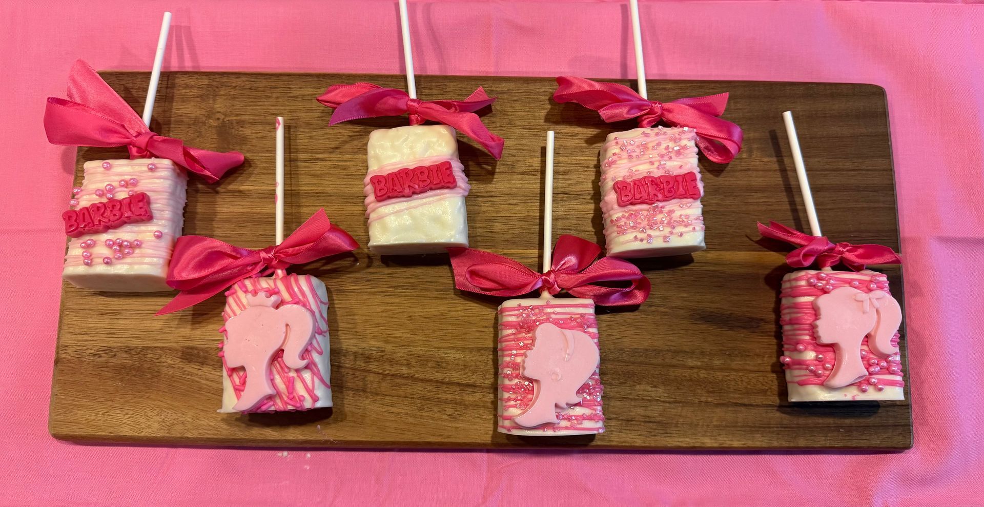 Marshmallow treats decorated with pink Barbie accents on a wooden board, tied with pink ribbons.