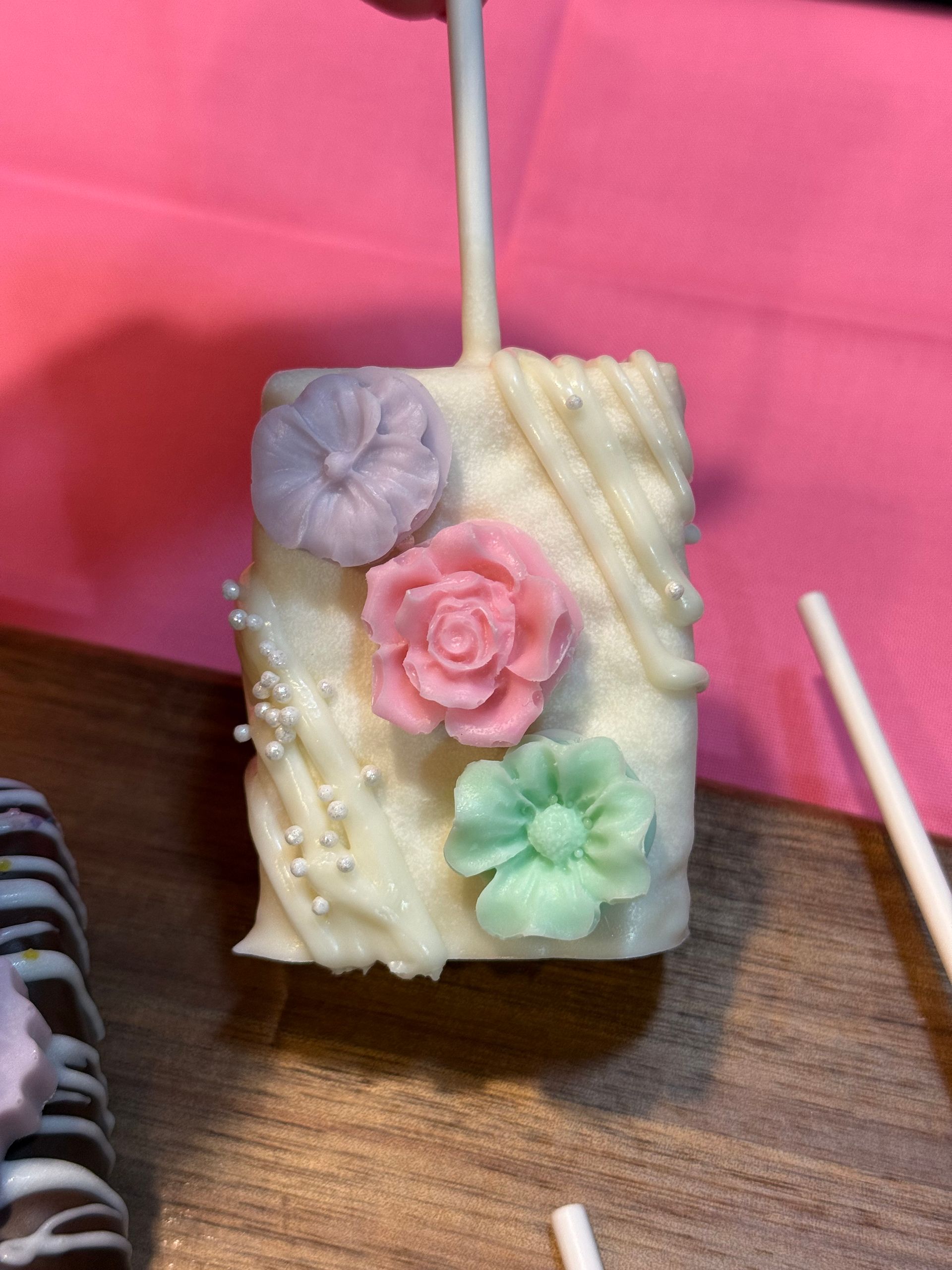 Marshmallow treat on a stick, coated in white chocolate, decorated with pastel flowers and sprinkles.