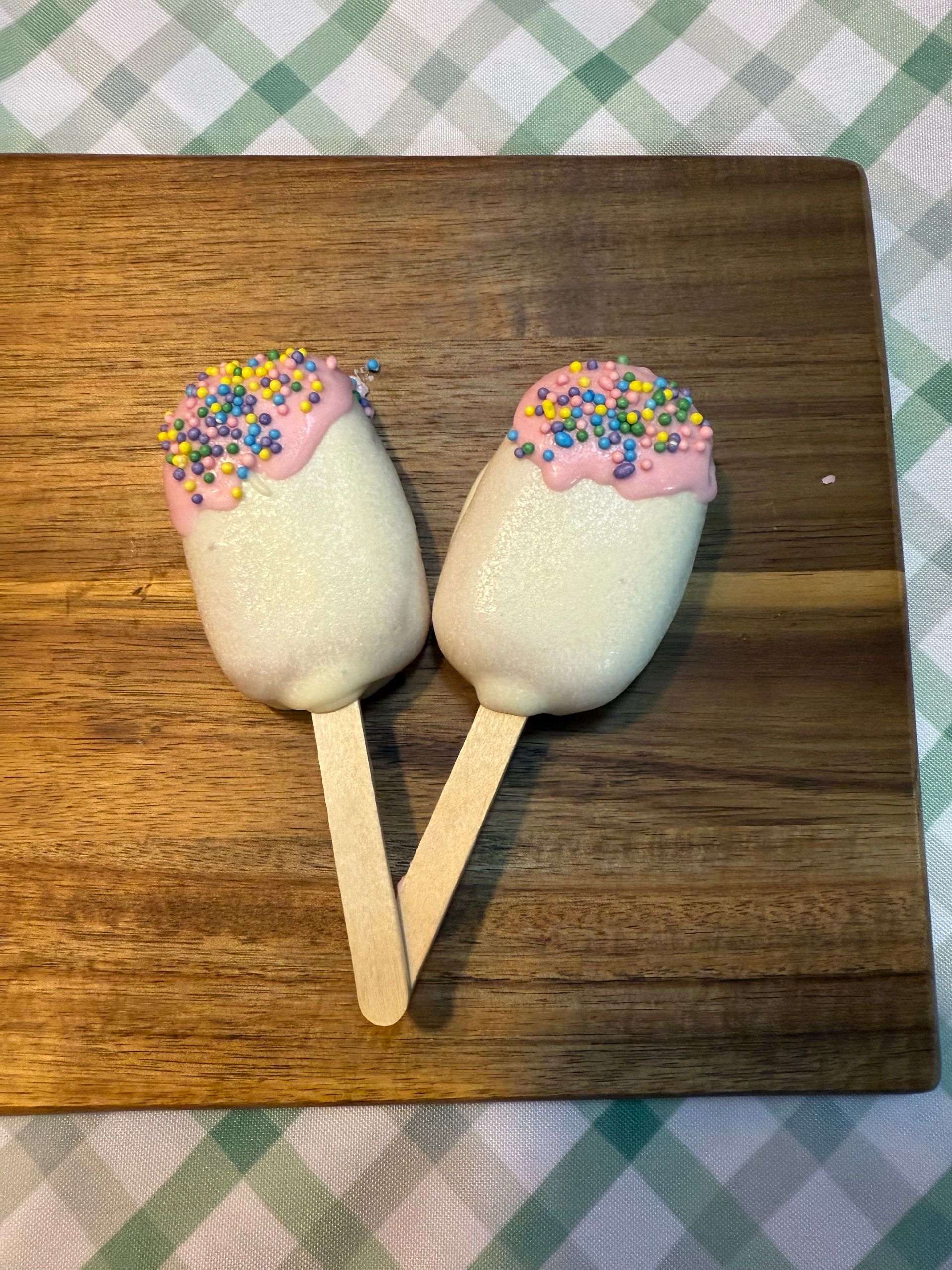 Two white-coated treats on wooden sticks, topped with pink frosting and sprinkles, on a wood board.