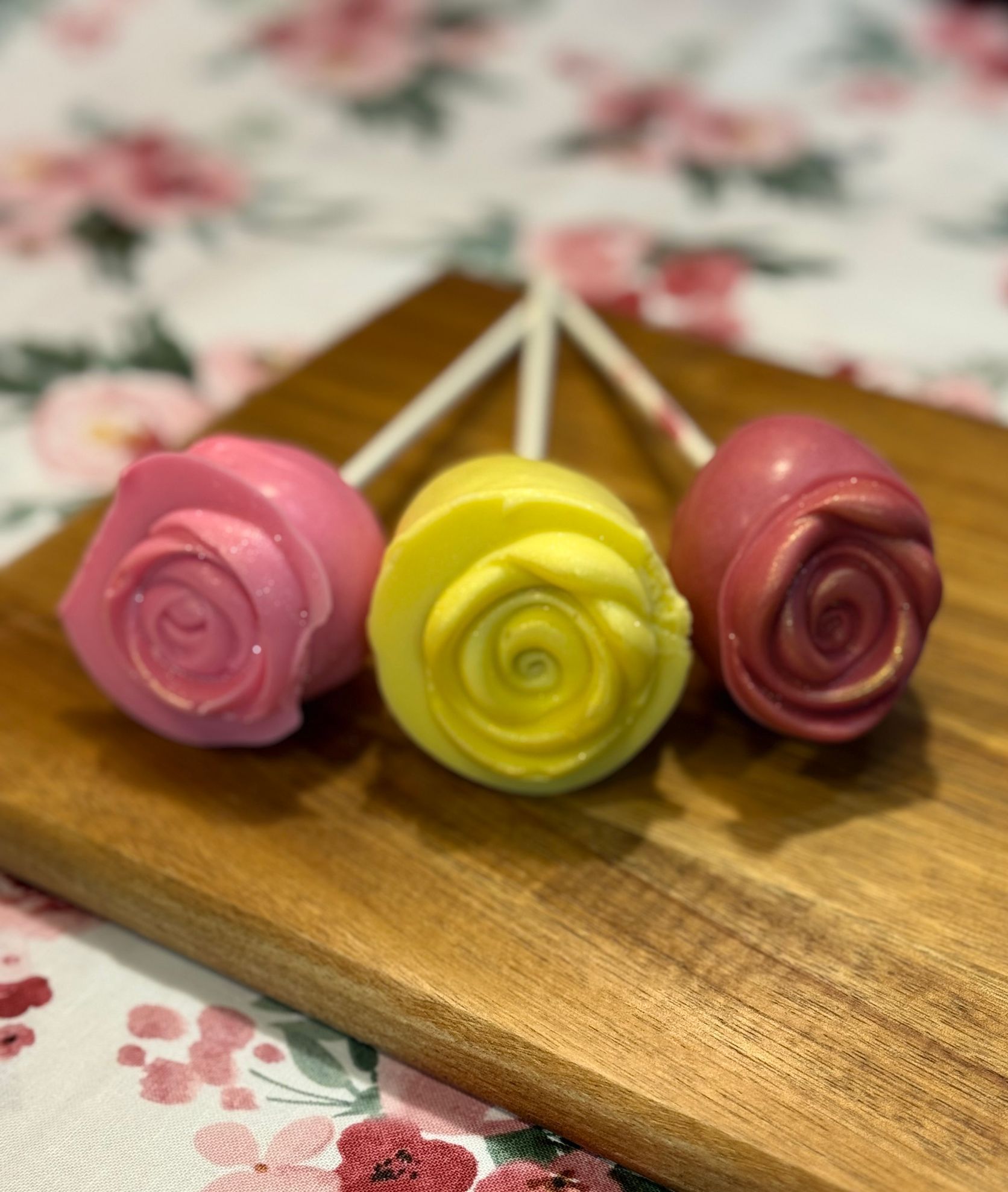 Three lollipops in the shape of roses are sitting on a wooden cutting board.