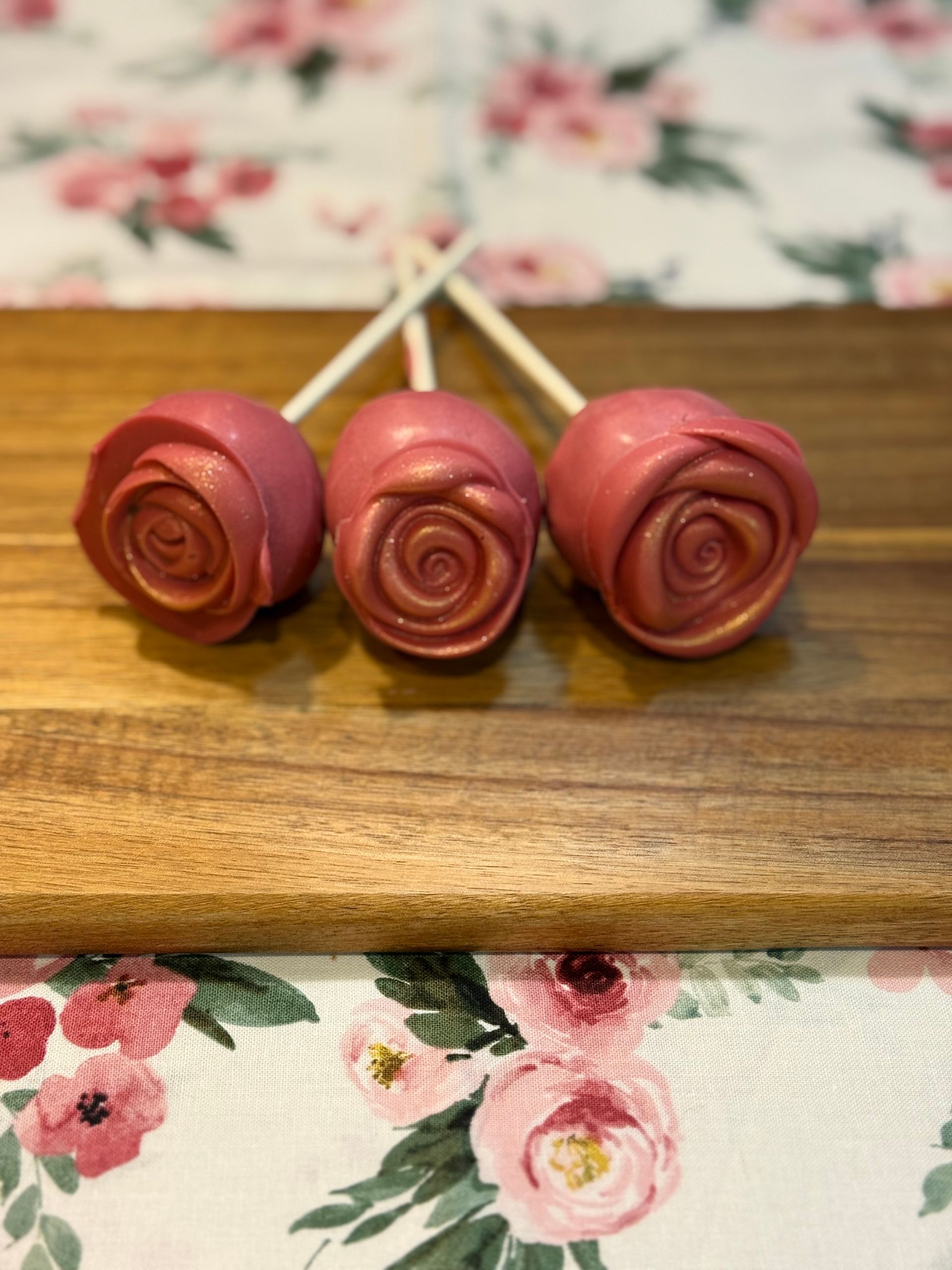 Three rose-shaped cake pops in metallic pink, displayed on a wooden board, floral tablecloth.