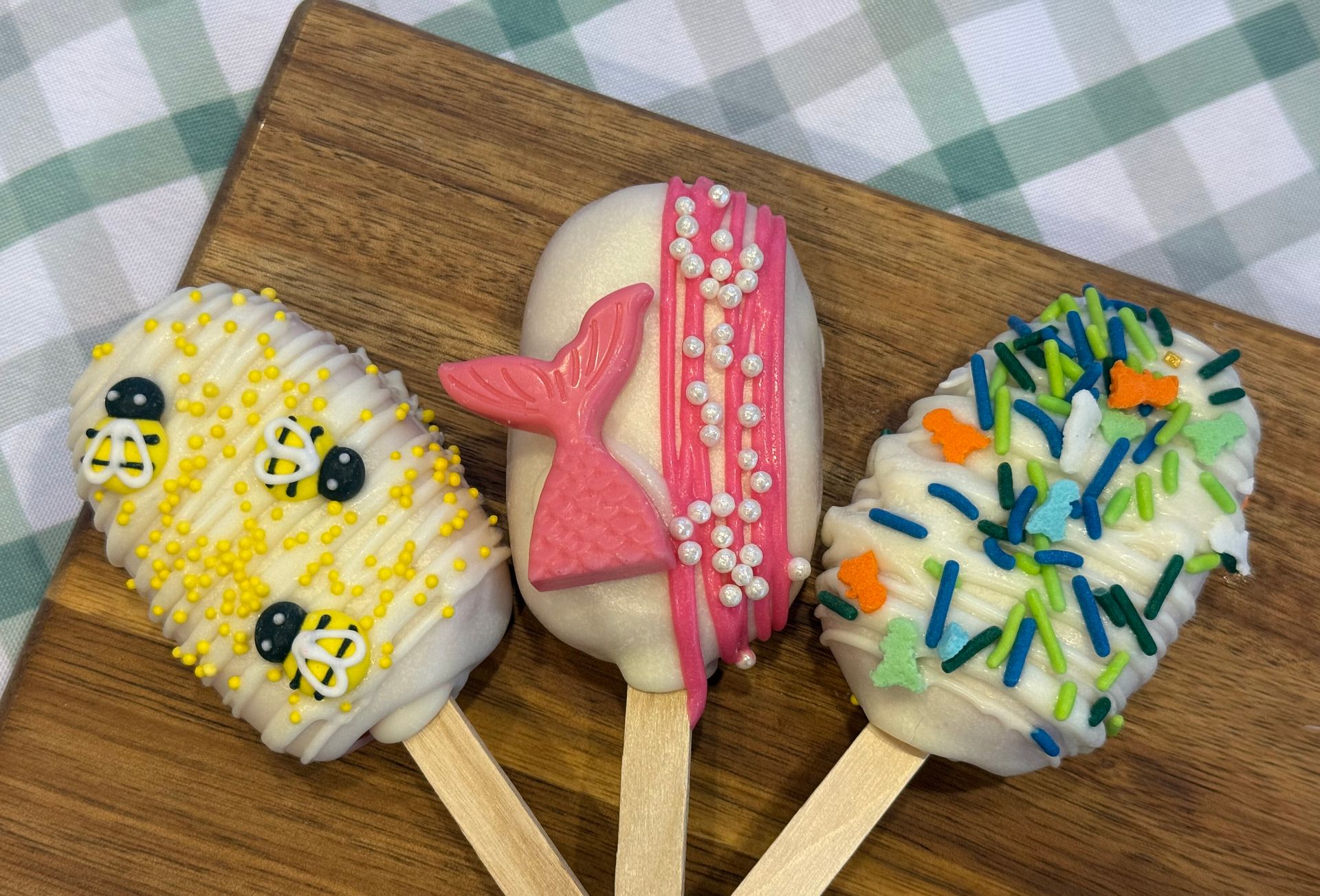Three cake pops: bee, mermaid tail, and colorful sprinkles on wooden board.