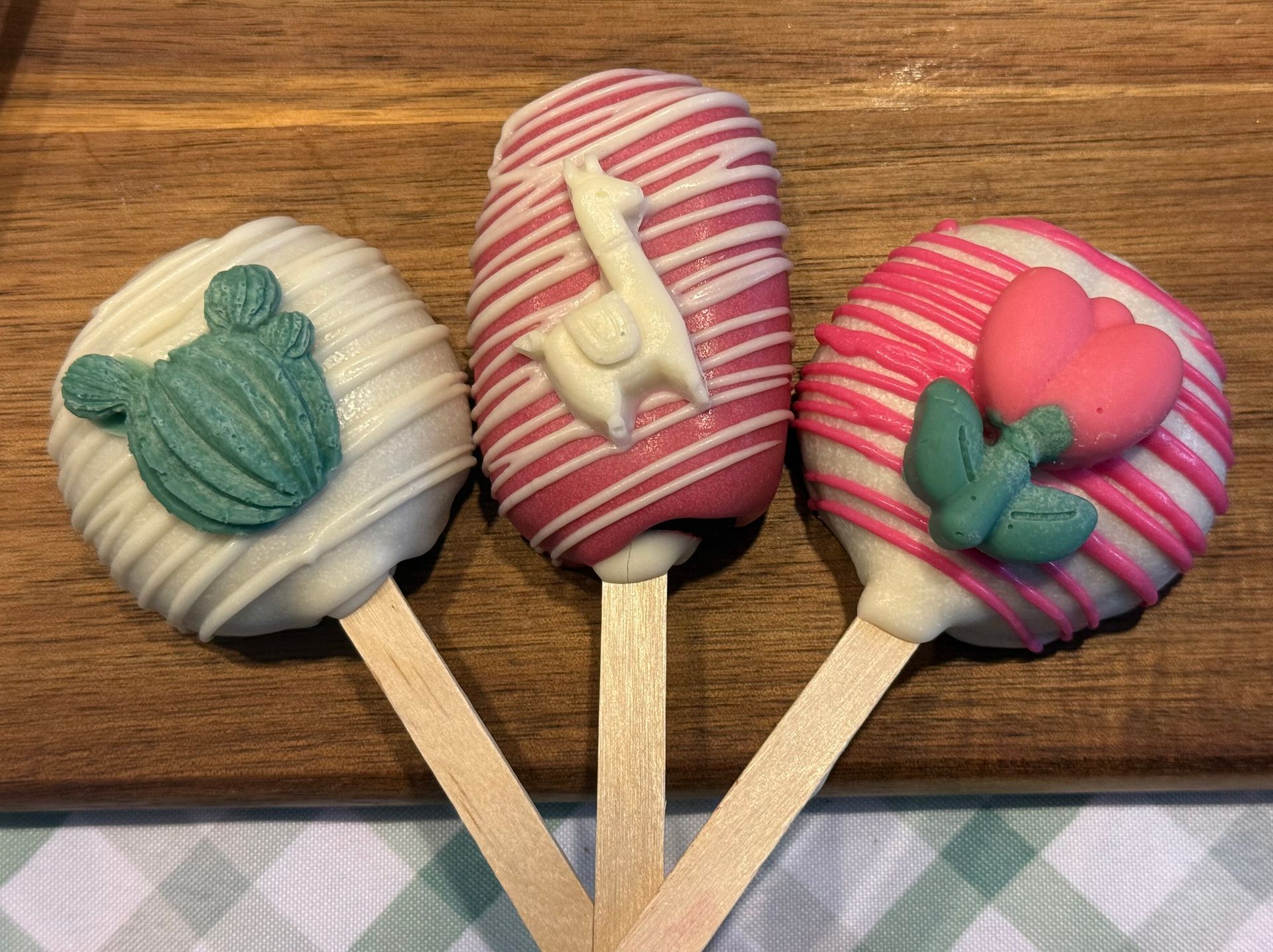 Three colorful cake pops on sticks: cactus, llama, and flower designs, on a wood surface.