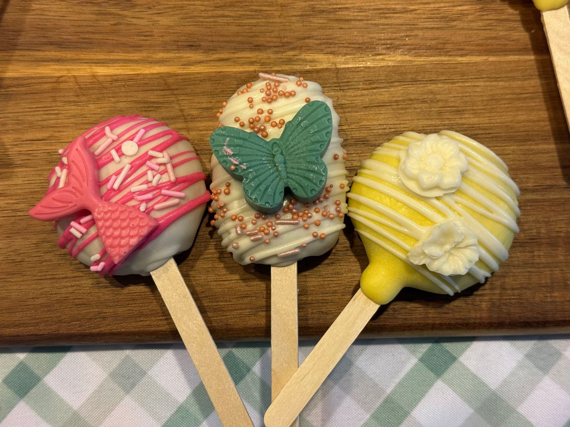 Three colorful cake pops on wooden sticks; a pink bow, a turquoise butterfly, and white flowers.