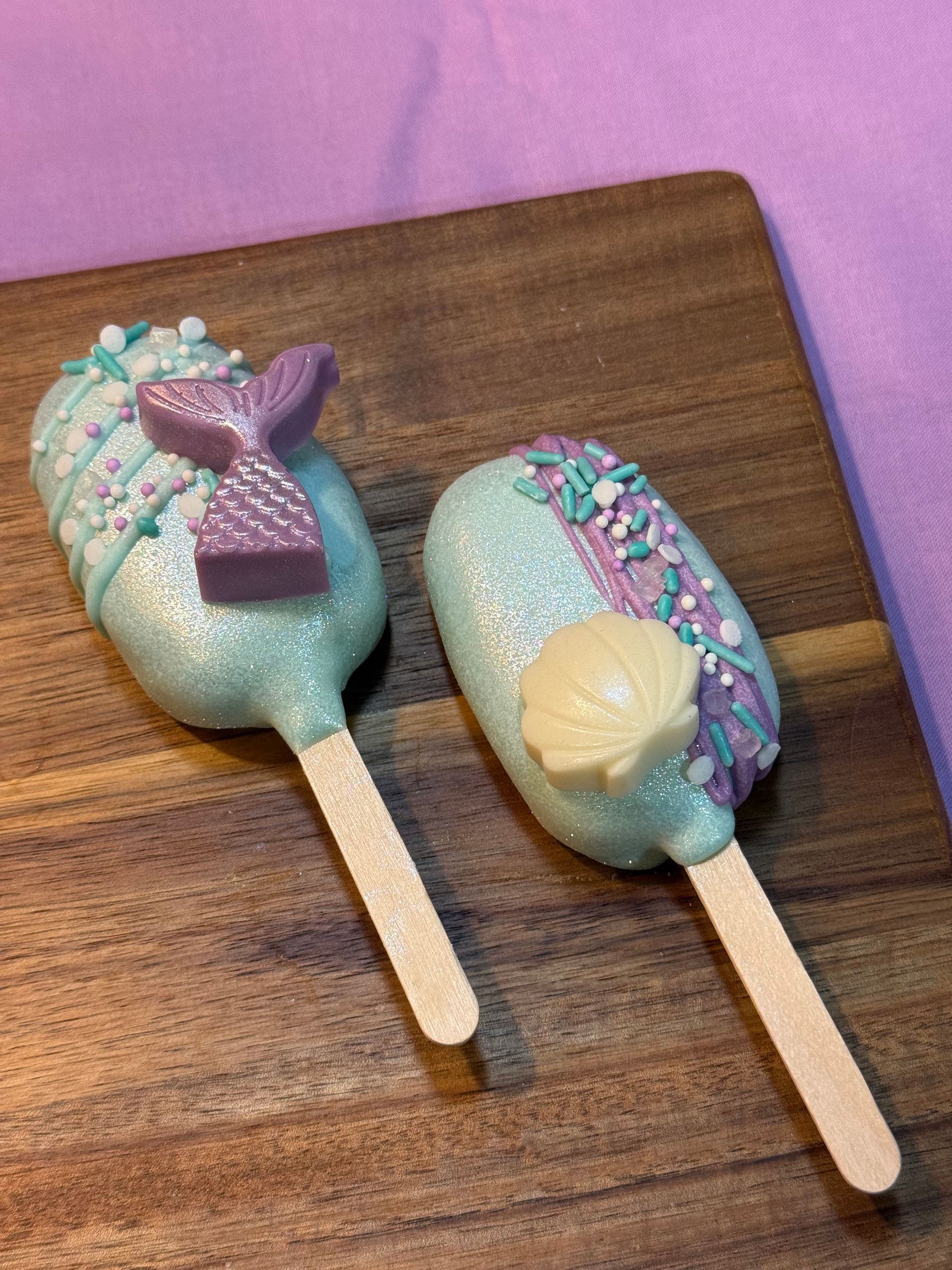 Two mermaid-themed cake pops: light blue with glitter, decorative sprinkles, and a seashell on a wooden surface.