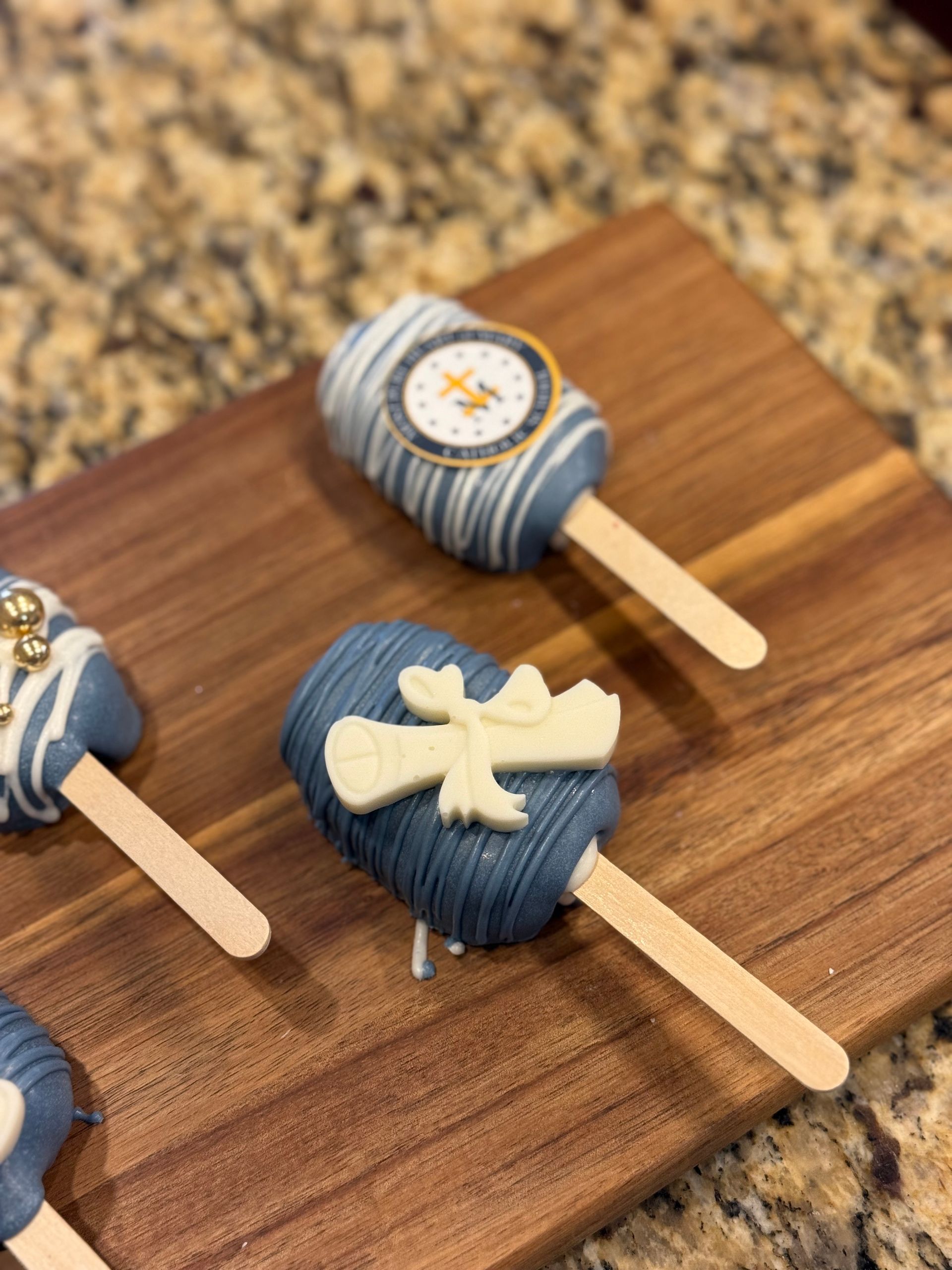 Cake pops on a wooden board. Blue and white with graduation-themed decorations, on popsicle sticks.