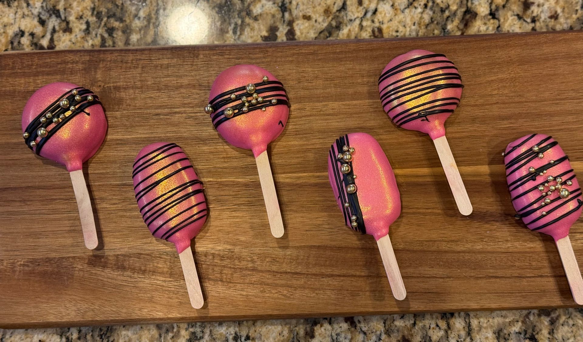 Pink cake pops on a wooden board, decorated with black stripes and sprinkles.