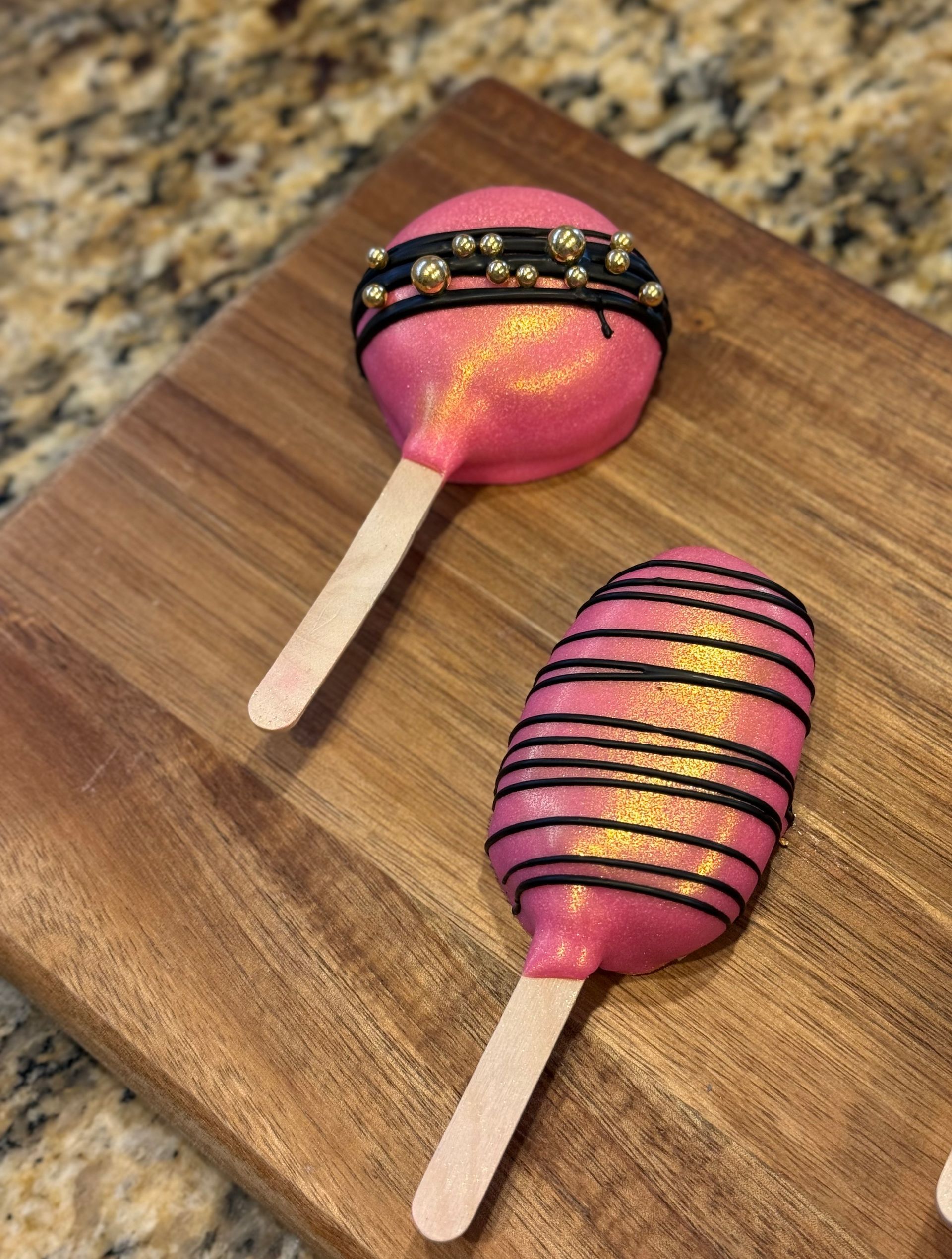 Two pink cake pops with decorative black lines and beads, on a wooden cutting board.