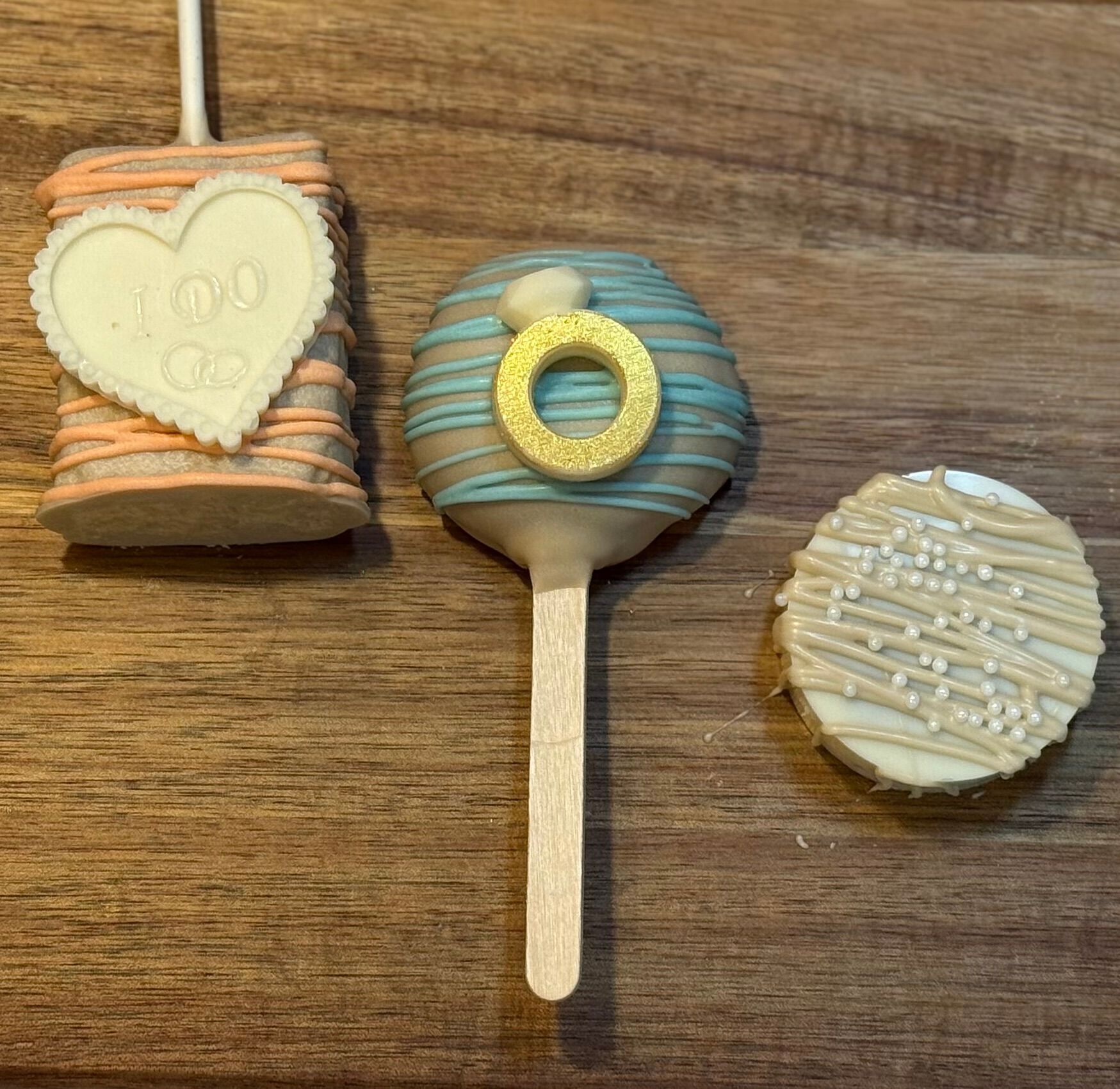Three engagement-themed treats: a heart-shaped marshmallow, a cake pop with a ring, and a decorated cookie on a wood table.