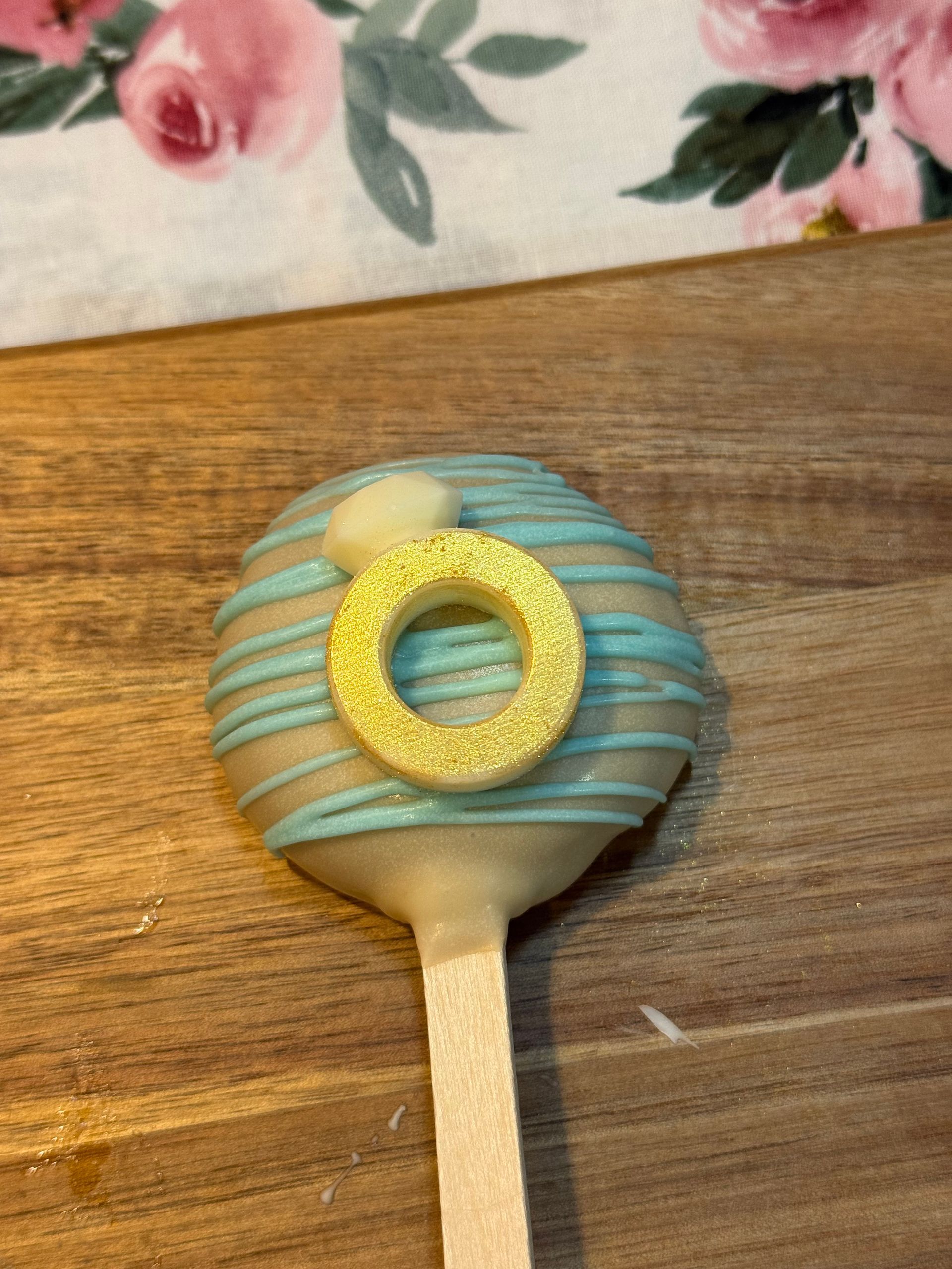 Cake pop decorated with a gold ring on blue frosting, on a wooden stick.