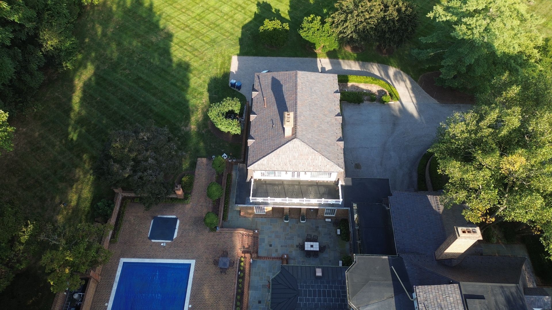 Aerial view of a home with a gray roof, a rectangular blue swimming pool, brick patio, and green trees.