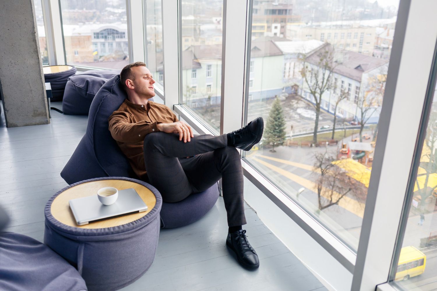 A man in a brown shirt and gray pants relaxes in a beanbag chair by a window, with a laptop and cup on a nearby table.