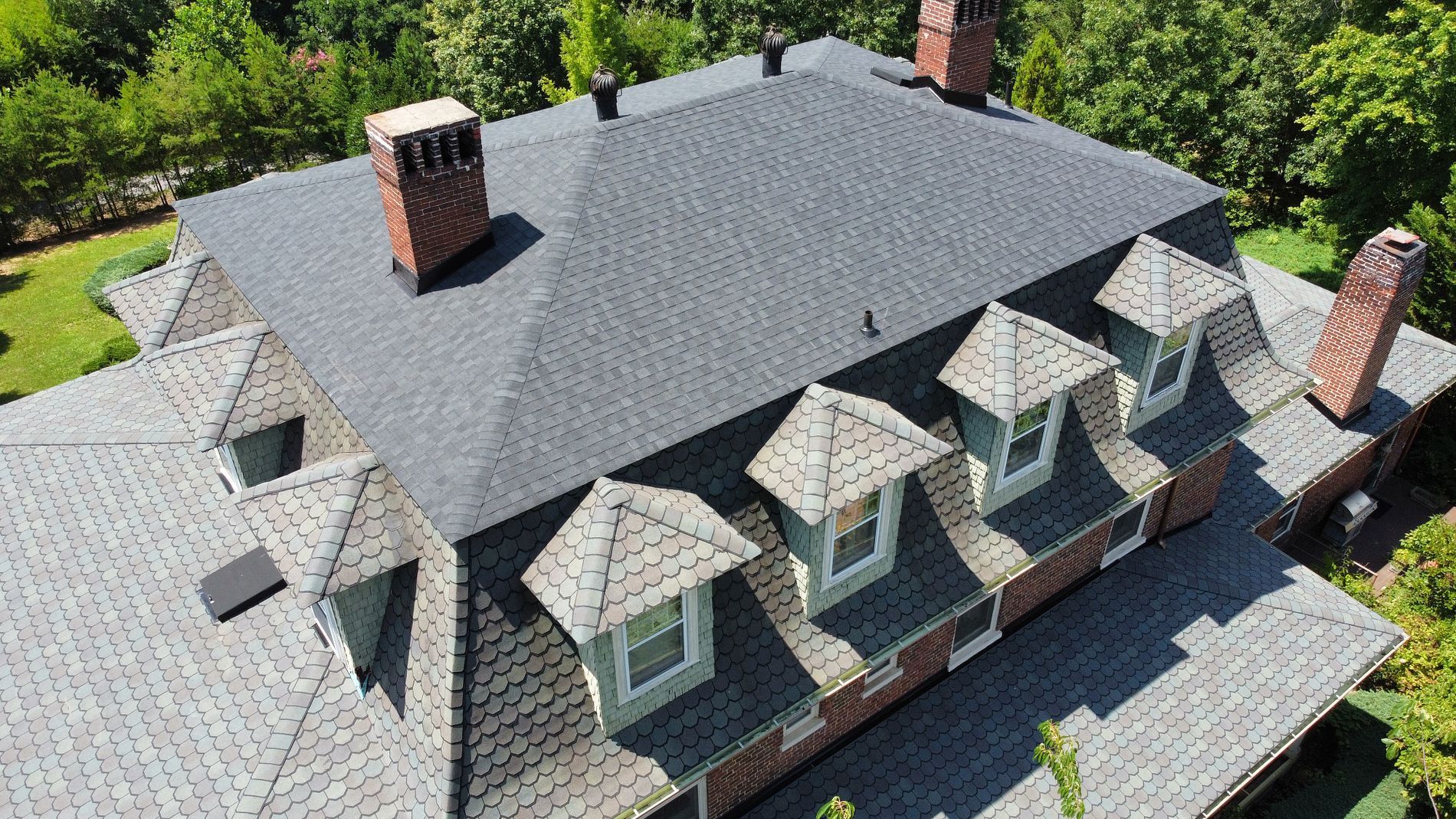 A high-angle aerial view of a dark shingled roof with several dormer windows and two brick chimneys on a sunny day.