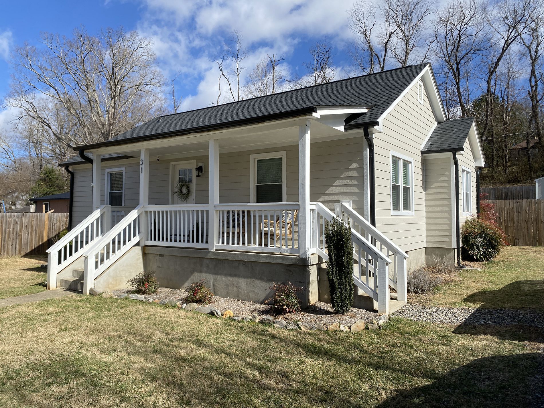 A one-story house with beige siding, a dark metal roof, and a white-railed front porch under a sunny blue sky.
