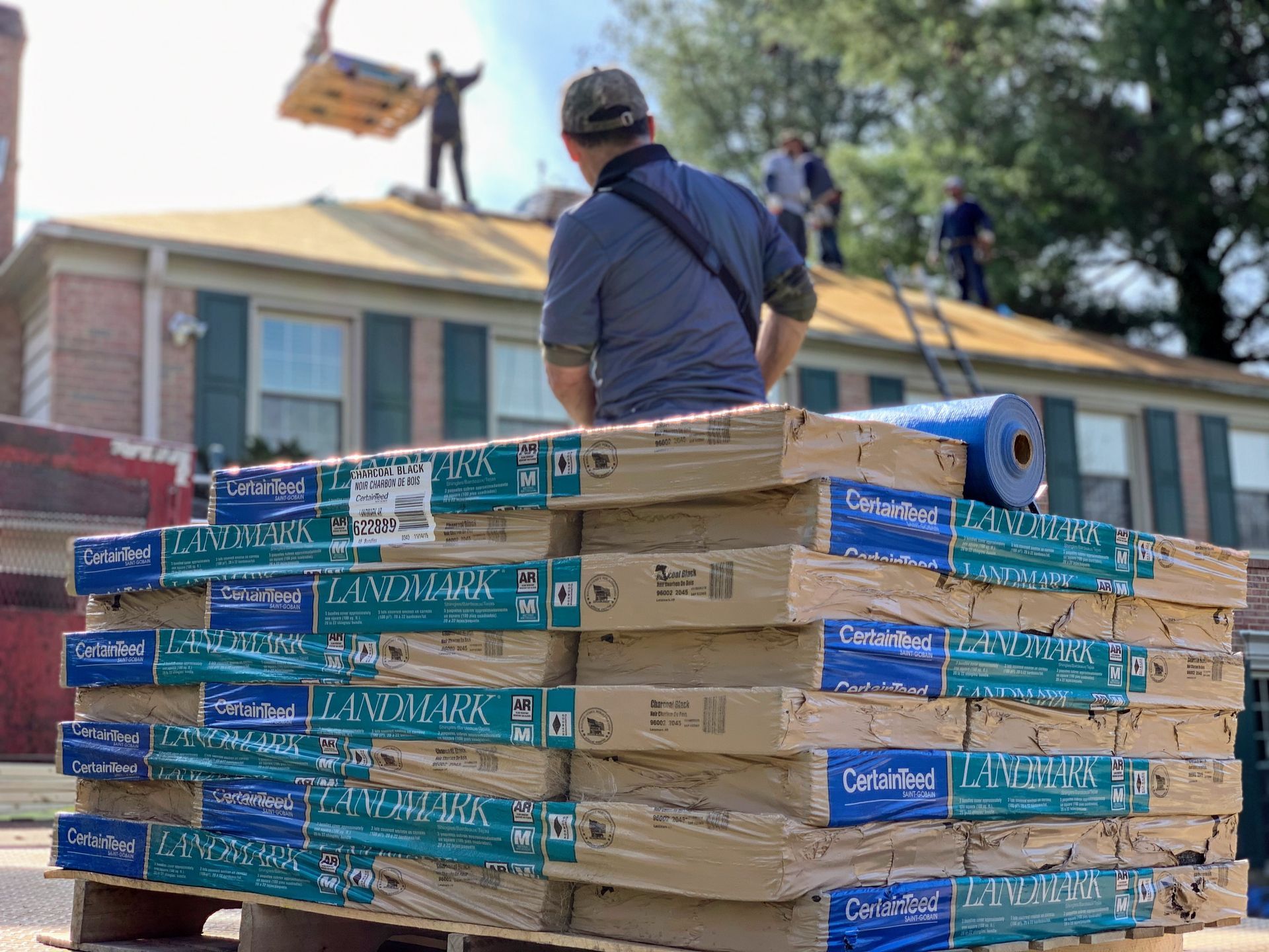 A worker stands next to a stack of roofing shingles as a crane lifts materials onto a house roof with others working above.
