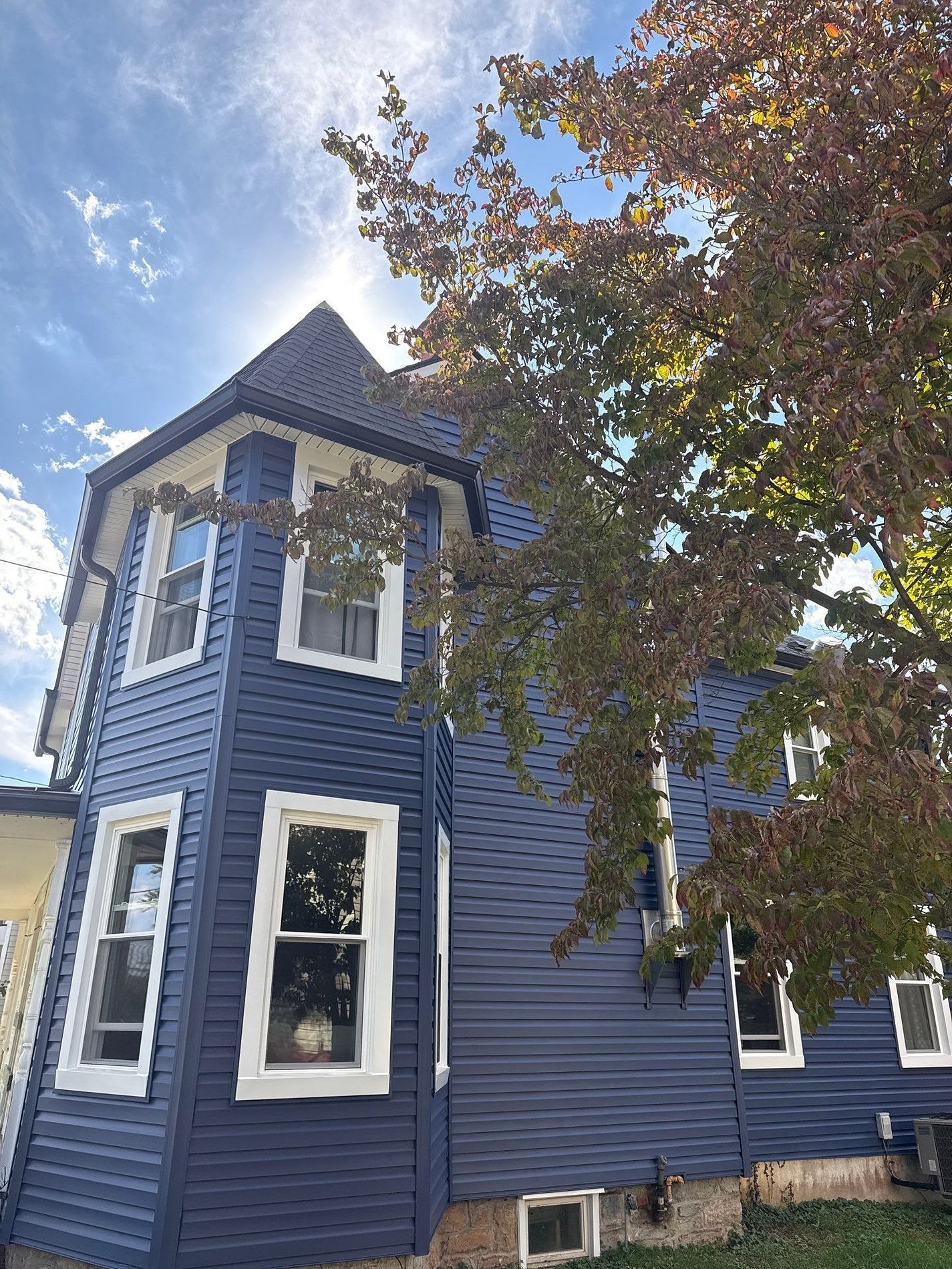 Blue Victorian house with a dark roof, framed by a leafy tree under a partly cloudy sky.
