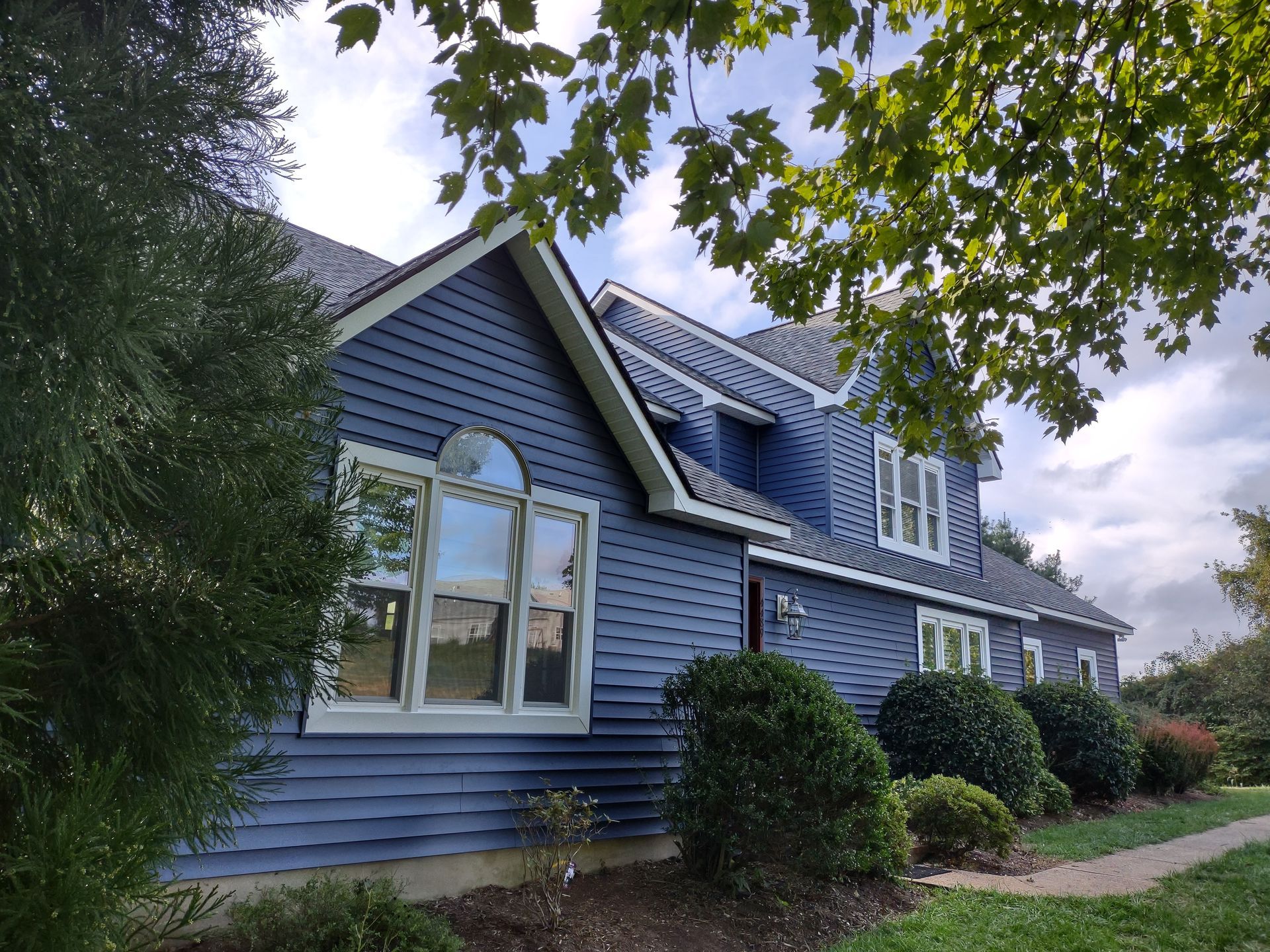 Blue-sided house with white trim and dark roof; surrounded by green bushes and trees under a cloudy sky.
