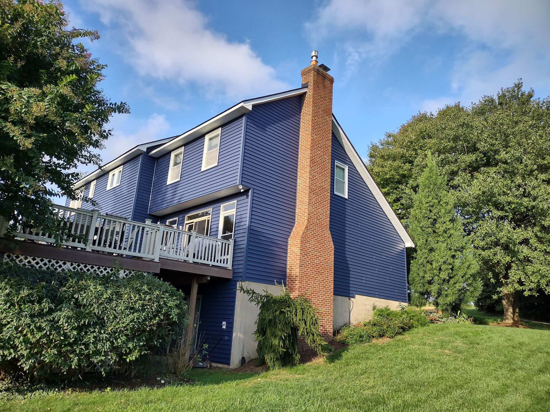 Blue-sided house with a brick chimney and deck, surrounded by green trees and grass under a blue sky.